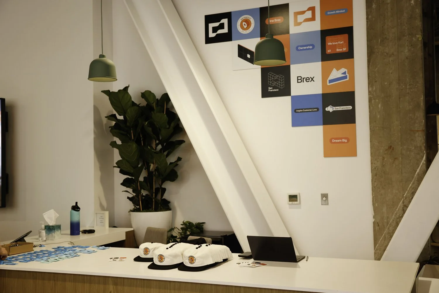 Indoor reception area with a large potted plant, white desk with hats and promotional materials, two green pendant lights, colorful wall art with words and icons, and a small digital display Brex headquarter in San Francisco, CA.