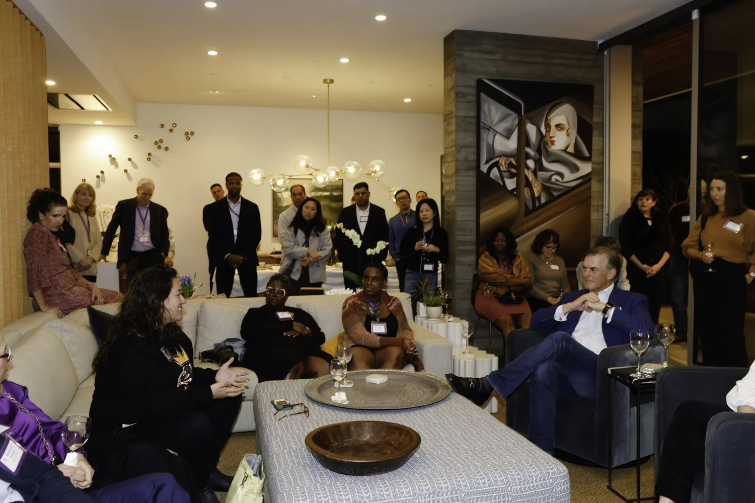 A diverse group of people gathered in a modern living room for a discussion or presentation, with some seated on couches and others standing, holding drinks, and listening attentively at Northwestern fundraiser in San Francisco, CA.