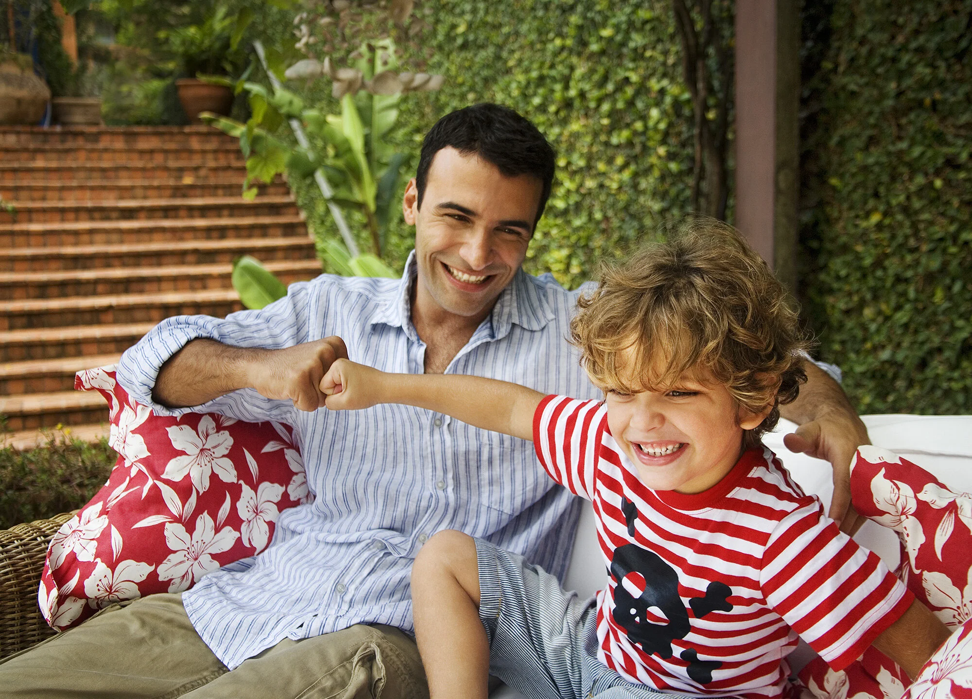 A Brazilian father and son playfully fist bump in an outdoor garden setting with brick stairs and green plants in the background in Rio de Janeiro, Brazil.