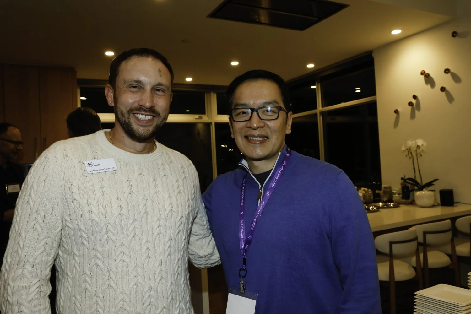 Two men standing and smiling at an indoor event. One has a beard and is wearing a white sweater; the other is wearing glasses and a blue shirt with an event lanyard Brex headquarter in San Francisco, CA.