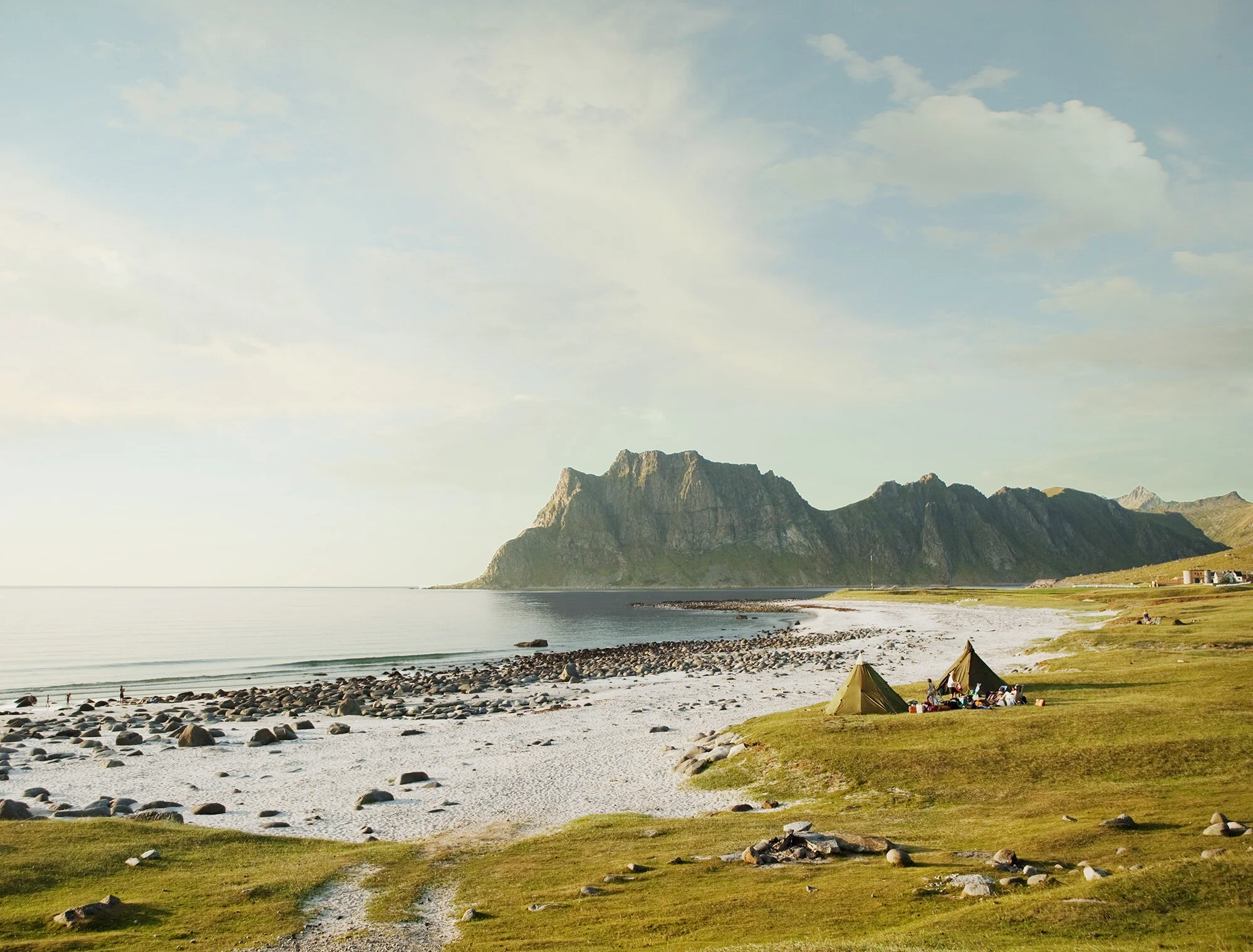 Scenic coastal landscape with two tents on grassy area, rocky beach, calm water, mountain range in the background, and a partly cloudy sky in Lofoten, Norway