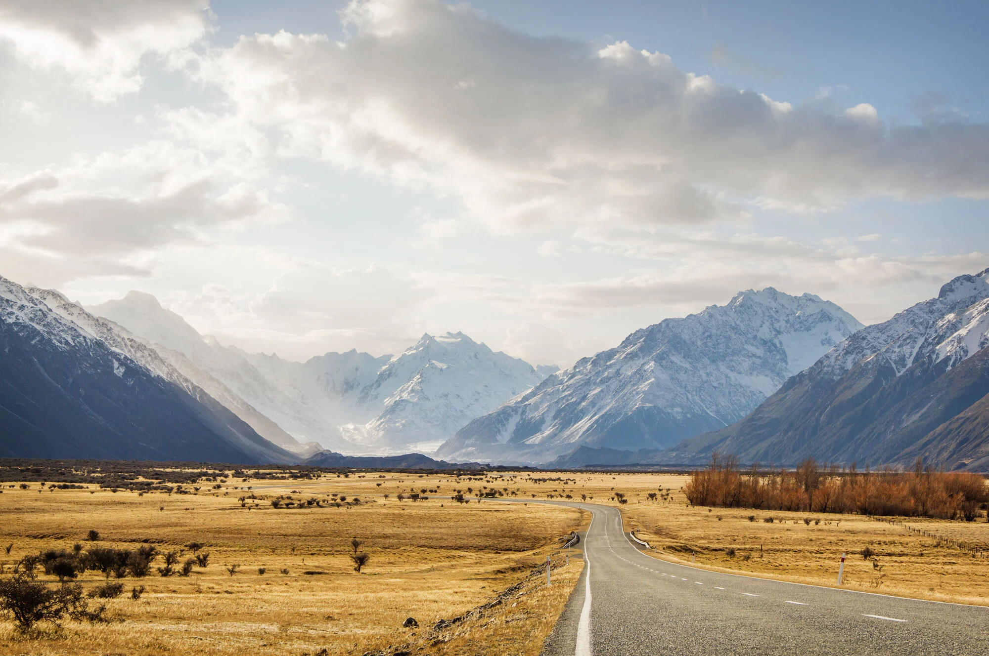 A winding road through a dry, grassy plain leading toward snow-capped mountains under a partly cloudy sky in New Zealand.