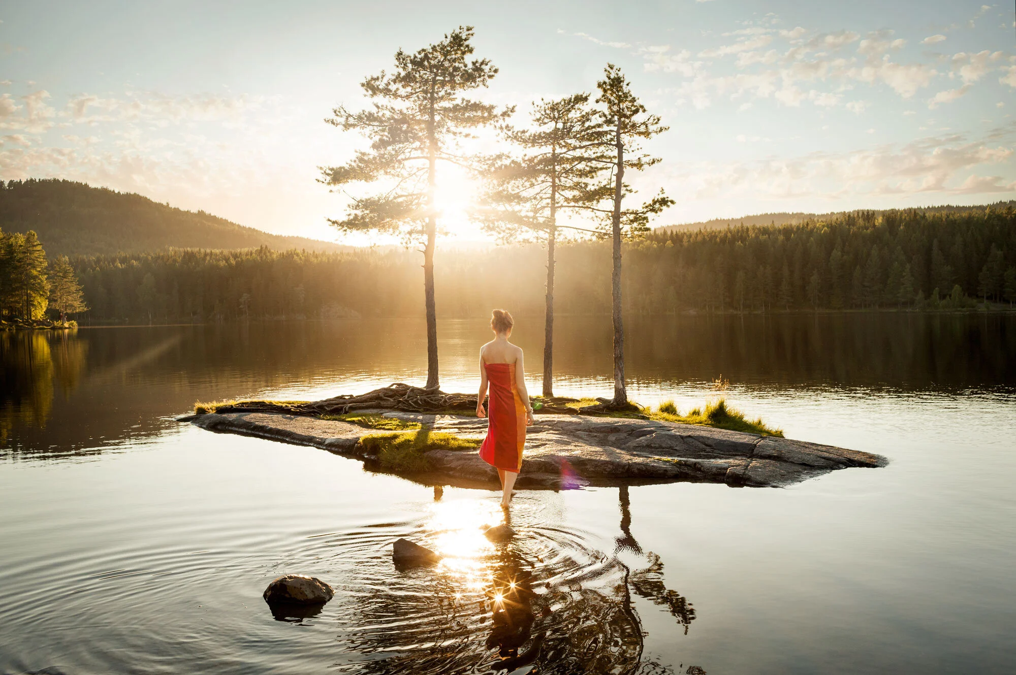 Woman in a red dress wading into a calm lake with a small island and three trees with backlit light, surrounded by a forested landscape during sunset at Oslo, Norway.