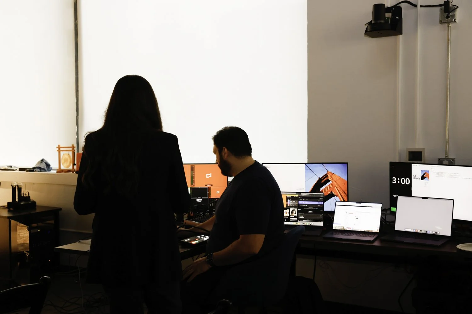 A woman and a man working in an office with multiple computer monitors, some displaying images of a building and others showing various applications; the woman stands while the man is seated in front of a device with controls Brex headquarter in San 