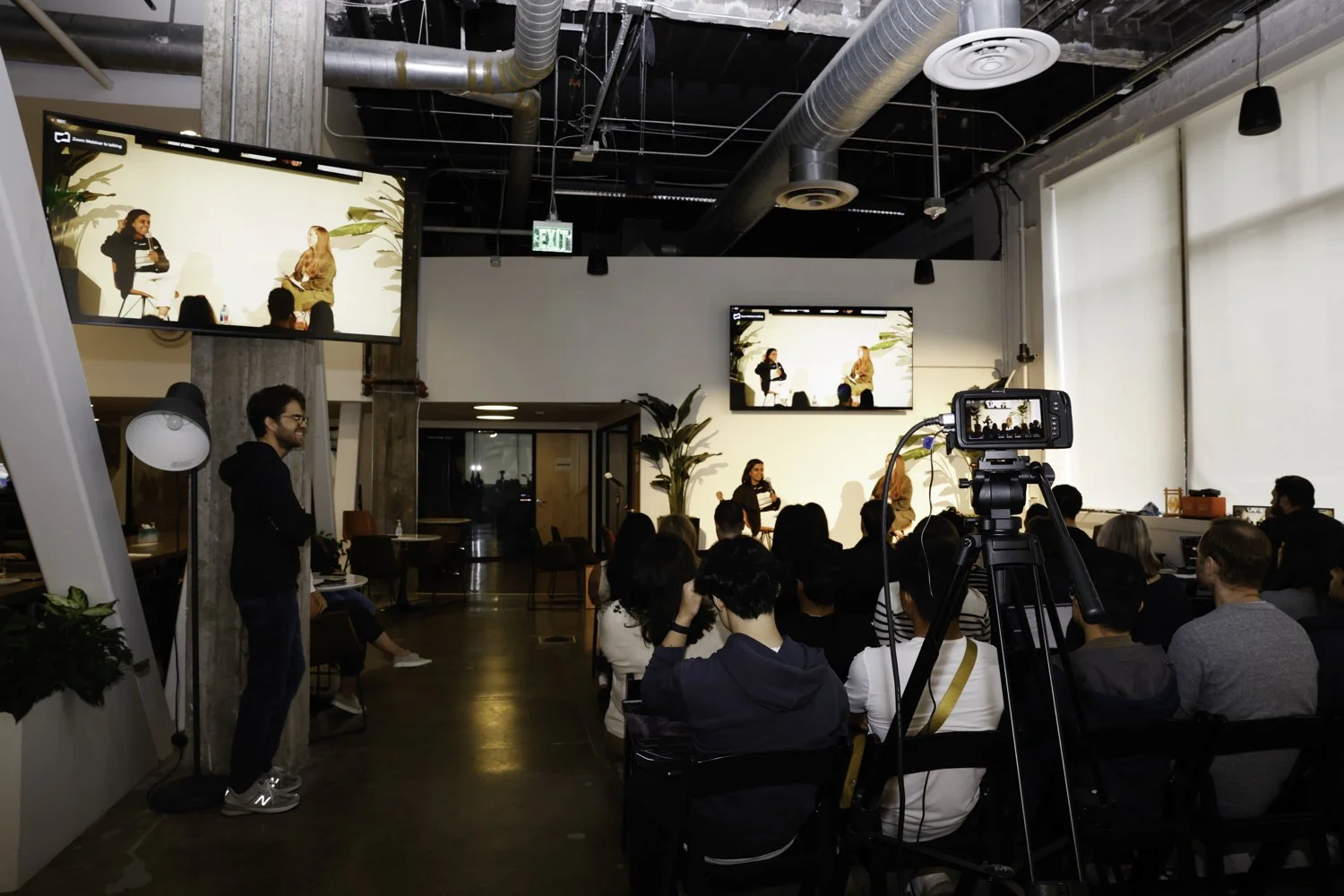 A panel discussion taking place in a modern, industrial-style venue with an audience watching two women on stage. Two large screens above the stage display the scene, and a camera on a tripod captures the event. The setting features exposed ductwork,