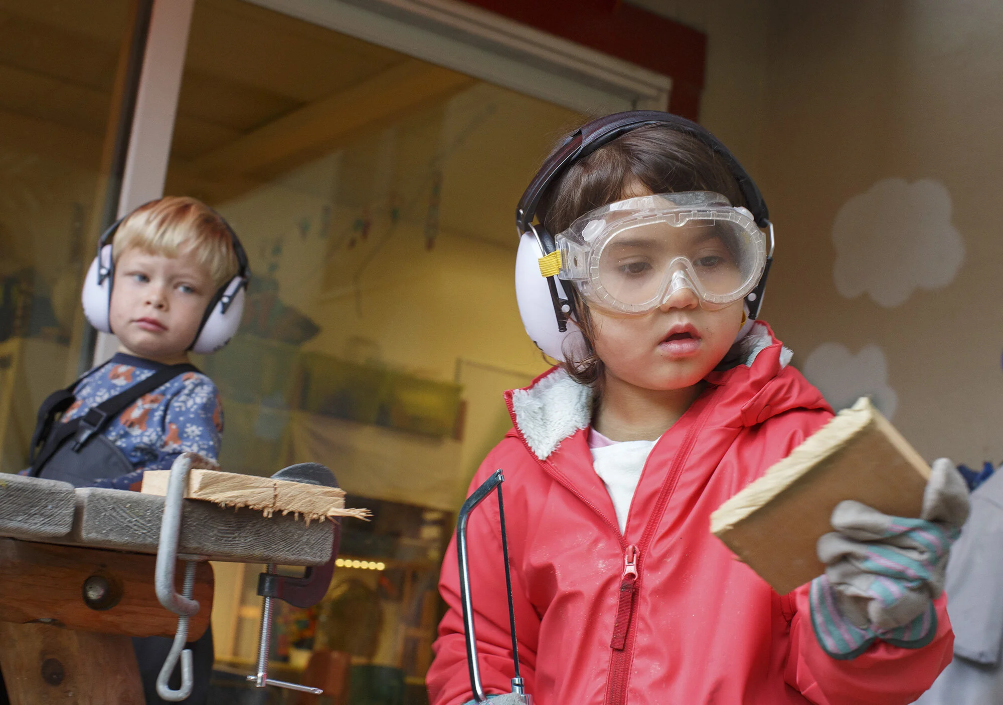 Two children wearing protective gear for woodworking; one child is holding a piece of wood and the other is observing, in a wood workshop setting The Scandinavian School in San Francisco, CA.
