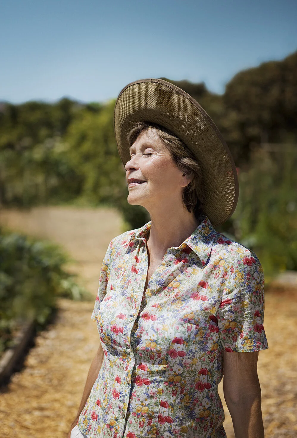 Senior white woman in a colorful floral shirt, wearing a wide-brimmed straw hat, enjoying the sunshine outdoors on a sunny day in San Francisco, CA.
