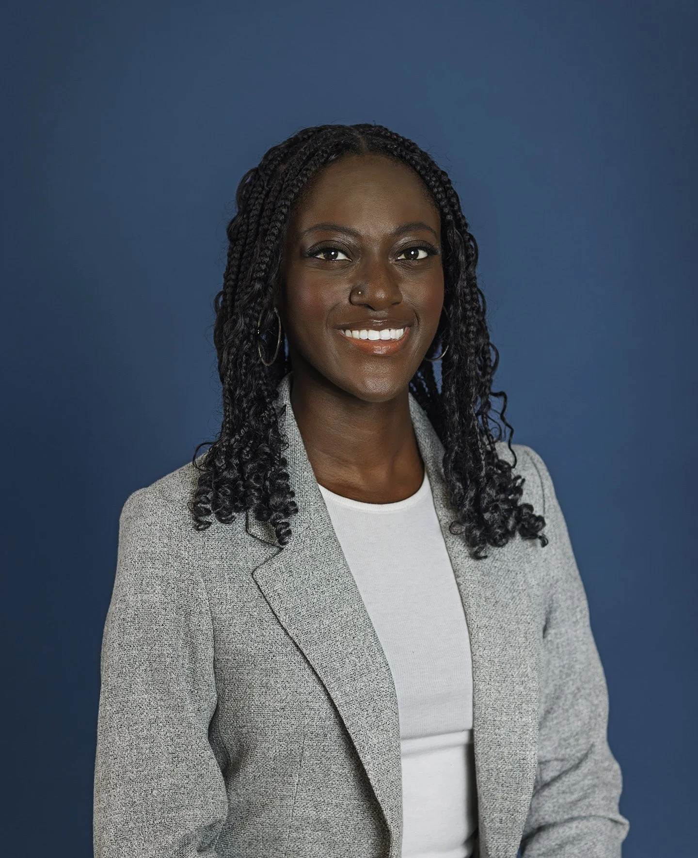A Black, young woman dressed in a white shirt and gray jacket with curly, long, black hair, smiling to the camera with beautiful white teeth against a blue backdrop in San Francisco, CA.