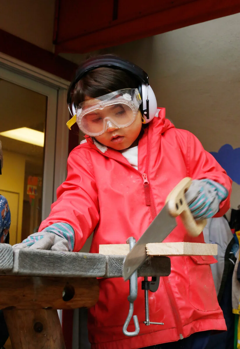 A young child wearing a red jacket, safety goggles, ear protection, and gloves is sawing a piece of wood held in place with a clamp on a wood workbench The Scandinavian School in San Francisco, CA.