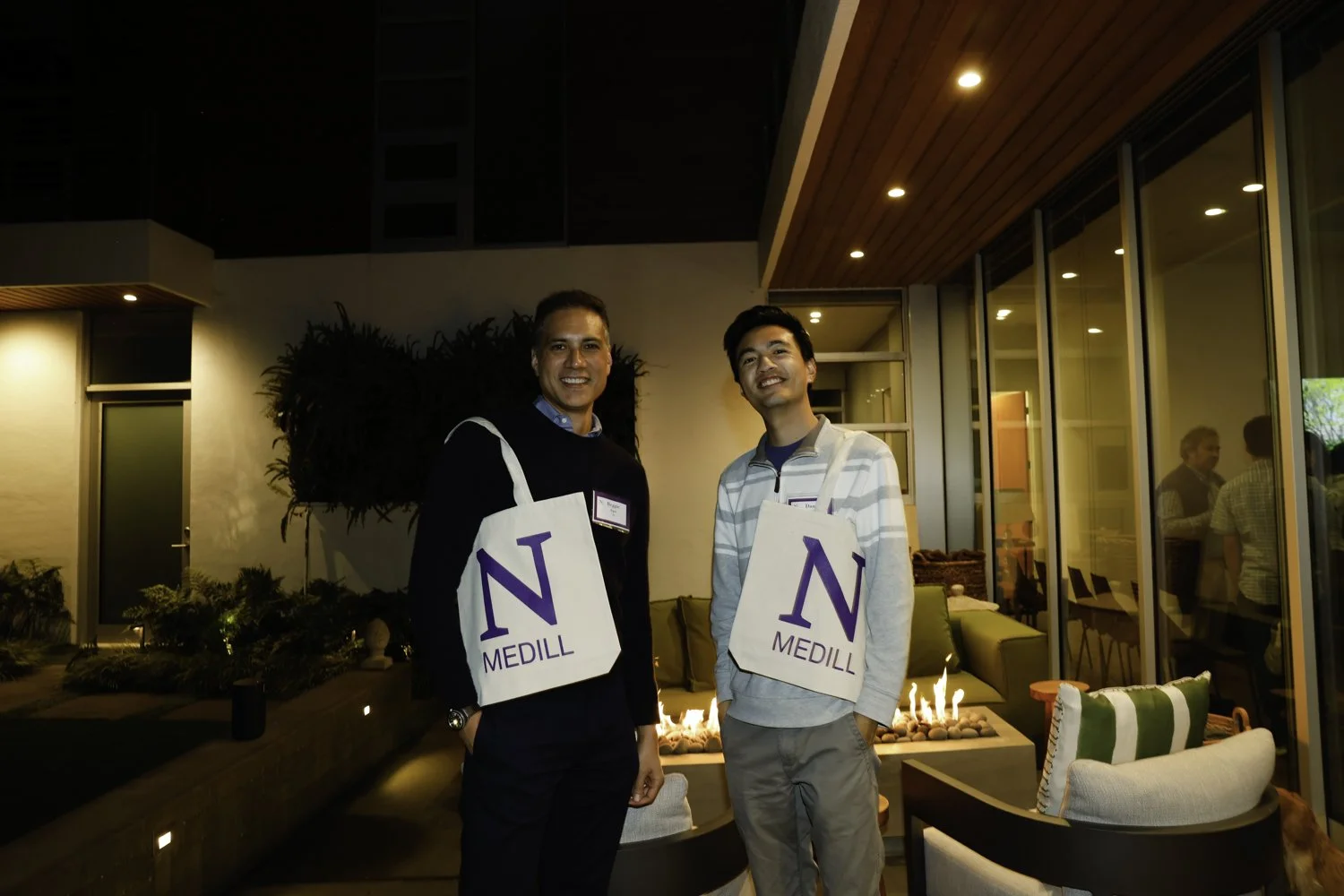 Two men smiling at an indoor evening event, each wearing a white tote bag with a purple N and the word 'MEDILL' on it, standing near a lit fireplace with seating, glass windows, and other people in the background at Northwestern fundraiser in San Fra