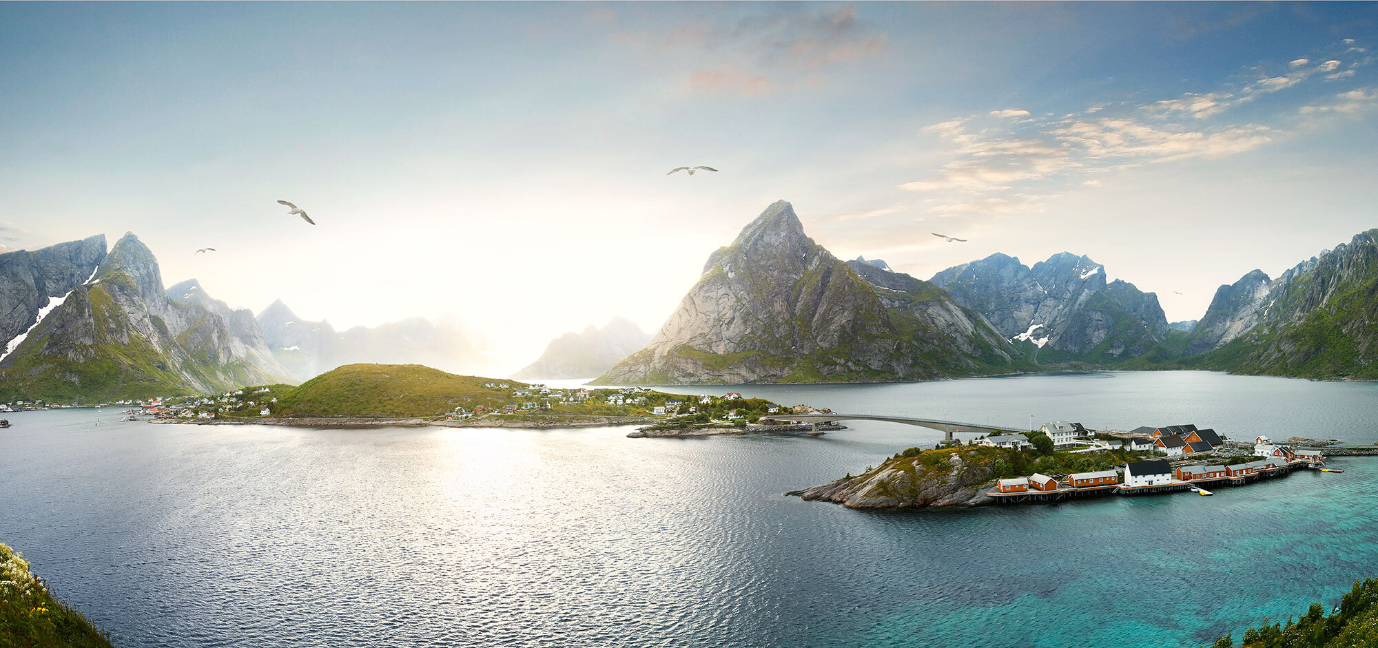 Scenic view of a fjord surrounded by tall, rugged mountains with some snow patches, a small island with fisherman's cabins, connected by a bridge, and a few birds flying in the sky in Lofoten, Norway.