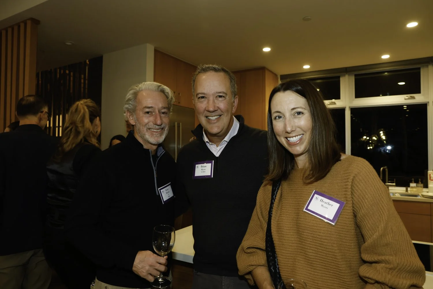 Three people smiling at an indoor social gathering, wearing name tags, with one person holding a wine glass, in a room with wooden cabinets and large windows Brex headquarter in San Francisco, CA.