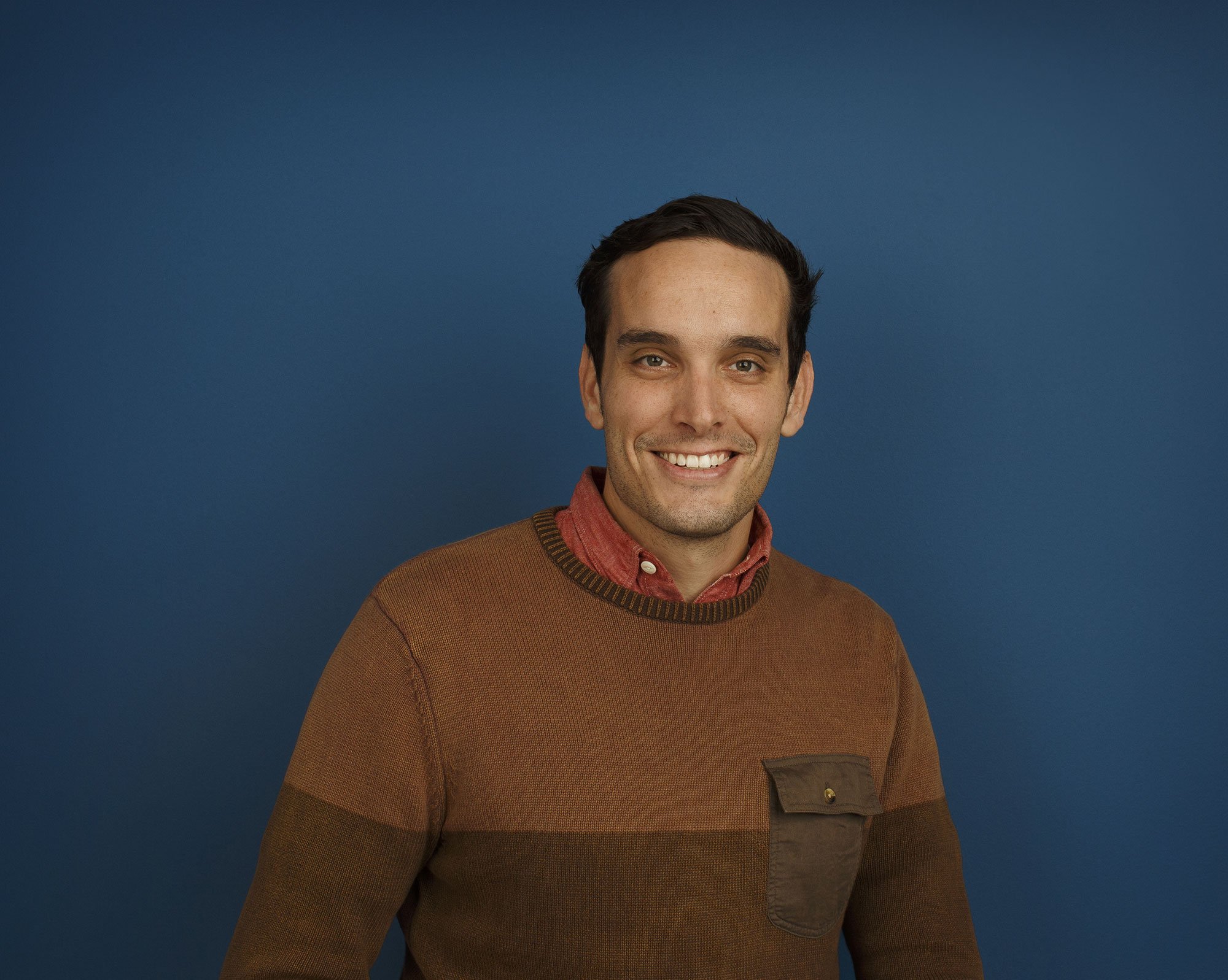 A young white man with dark, short hair and light skin, smiling, wearing a brown sweater with a chest pocket and a red collared shirt underneath, standing against a solid blue background in an architectural office in San Francisco, CA.