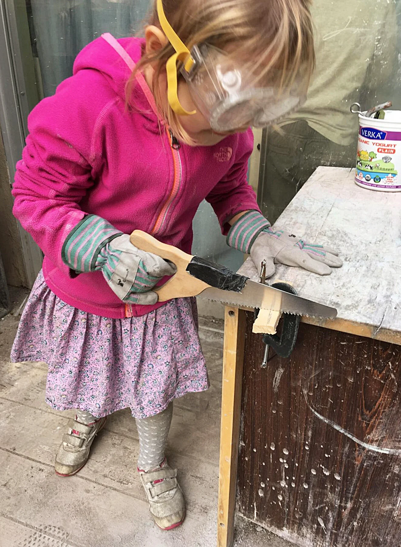 A young girl wearing safety goggles, a pink jacket, and gloves, using a saw to cut a piece of wood clamped to a workbench in a woodworking or crafting area at the The Scandinavian School in San Francisco, CA.