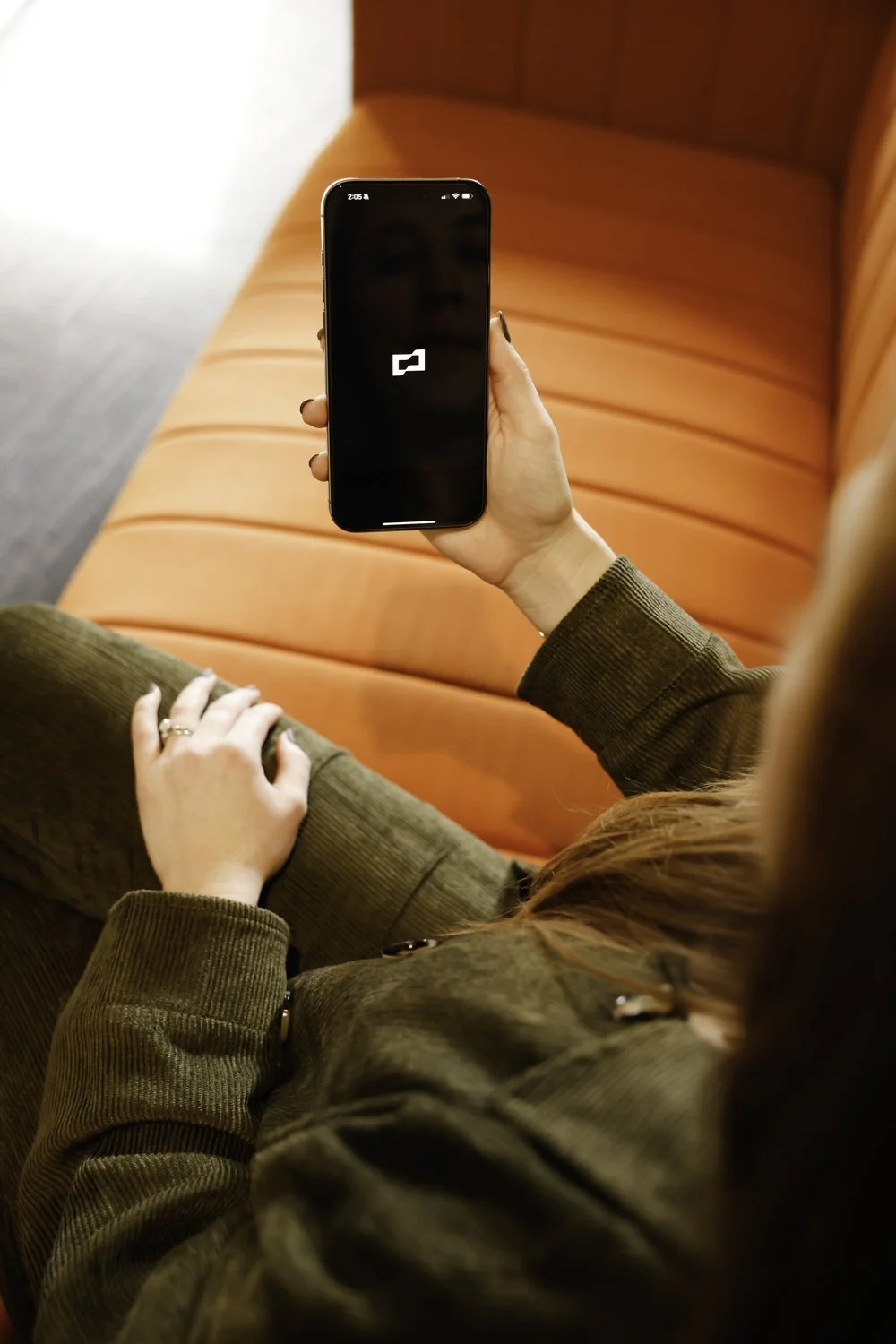 A woman with red hair holding a smartphone displaying an Brex logo, sitting on an orange sofa at Brex Headquarter in San Francisco, CA.