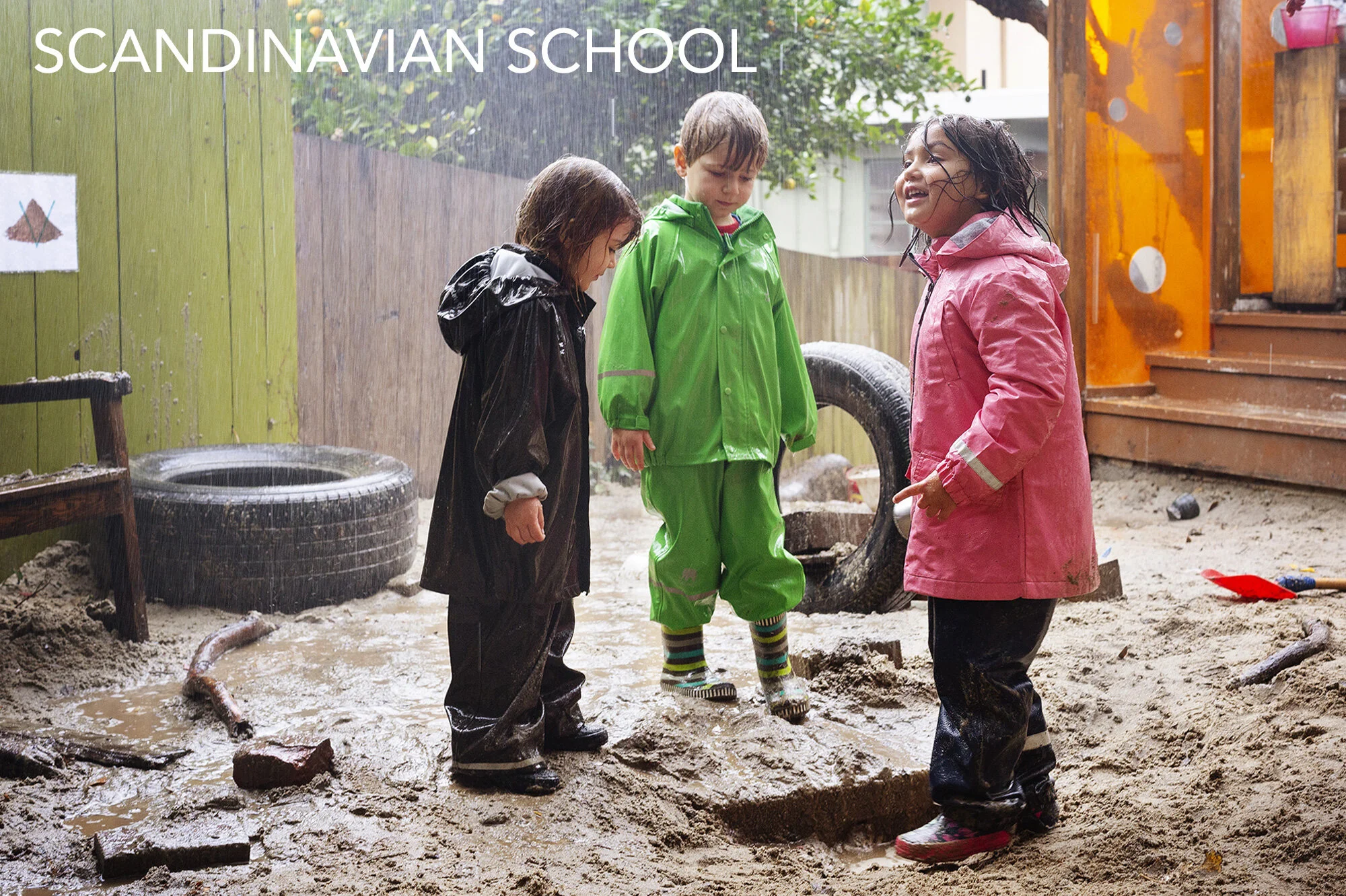 Three children in colorful raincoats and rainboots playing in a muddy sandbox during rainy weather at The Scandinavian School, San Francisco, CA.