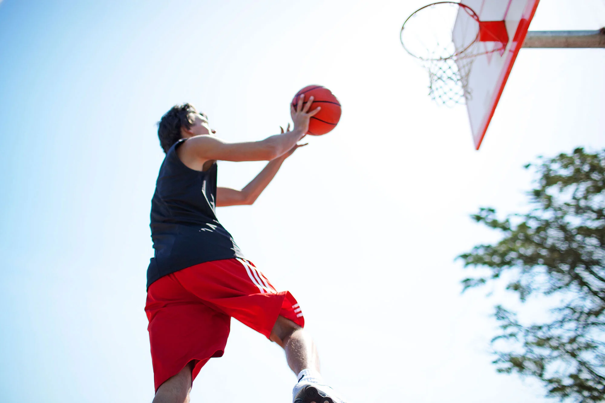 Young Latino man playing basketball outdoors in red shorts and a black shirt, jumping towards the hoop to shoot the ball on a sunny day in San Francisco, CA.