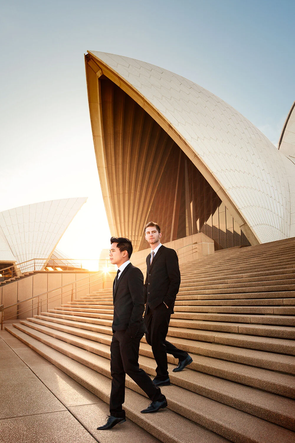 Two men, a couple, in black suits walking down the stairs in front of the Sydney Opera House at sunset in Sydney, Australia.