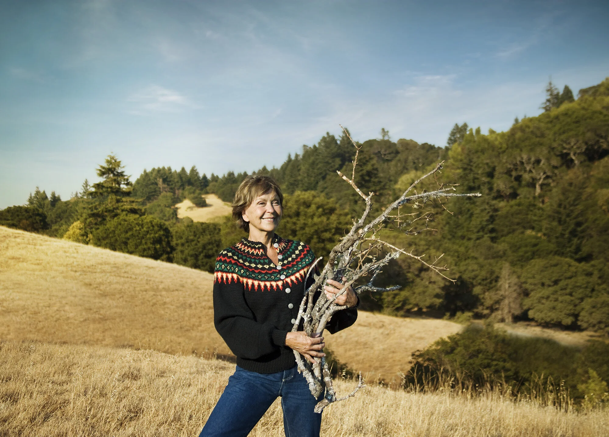A white woman standing in a yellow field holding a large, leafless branch or tree trunk. She is smiling and wearing a colorful knitted sweater and blue jeans. The background features hills, trees, and a bright blue sky in San Francisco, CA.