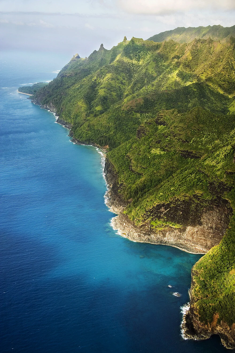 Aerial view of lush green cliffs along a vibrant blue ocean coastline in Kaua'i, Hawai'i.