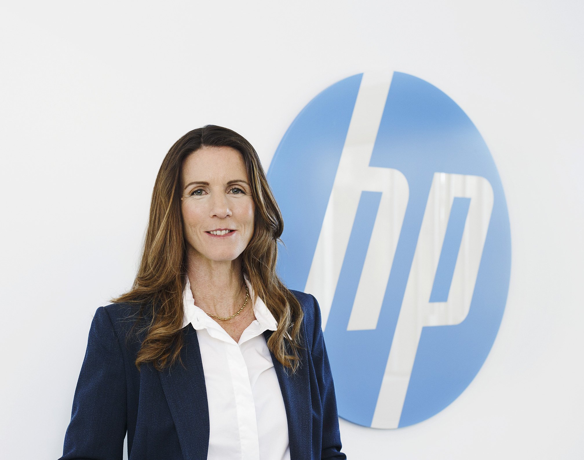 White woman CMO wearing a white shirt and blue blazer, shoulder length hair and a necklace, standing in front of a large Hewlett Packard logo at Hewlett Packard's office in Palo Alto, CA.