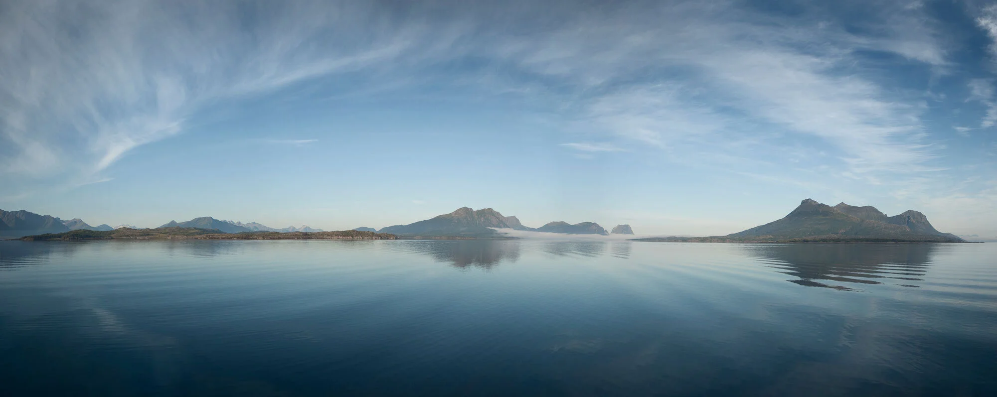 A panoramic view of calm body of water reflecting distant mountains and a partly cloudy sky in Lofoten, Norway.