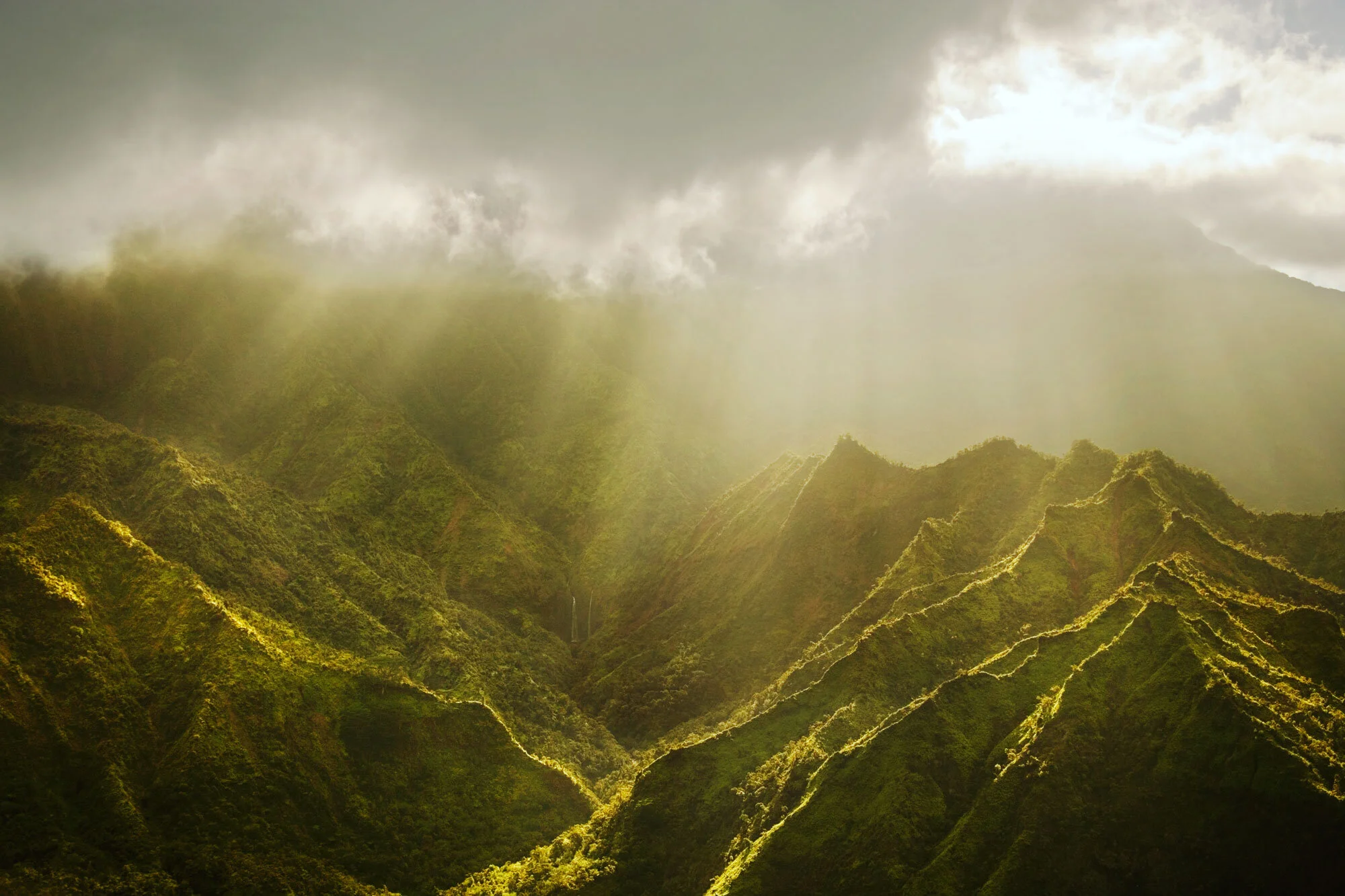 Sun rays shining through clouds over lush green mountains in an arial view from a helicopter in Kaua'i, Hawai'i.