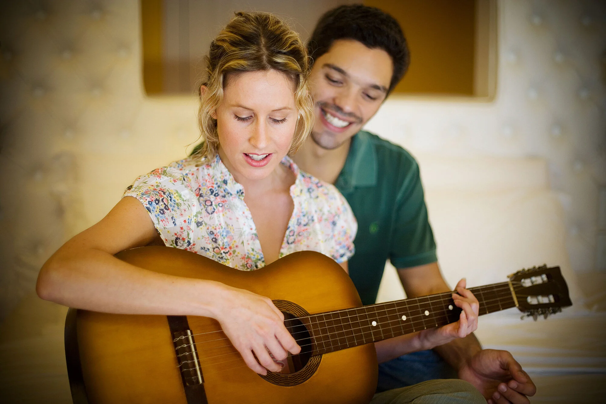 A Brazilian woman playing an acoustic guitar while a Brazilian man watches and smiles in a cozy, well-lit room at Boutique hotel in Rio de Janeiro, Brazil.