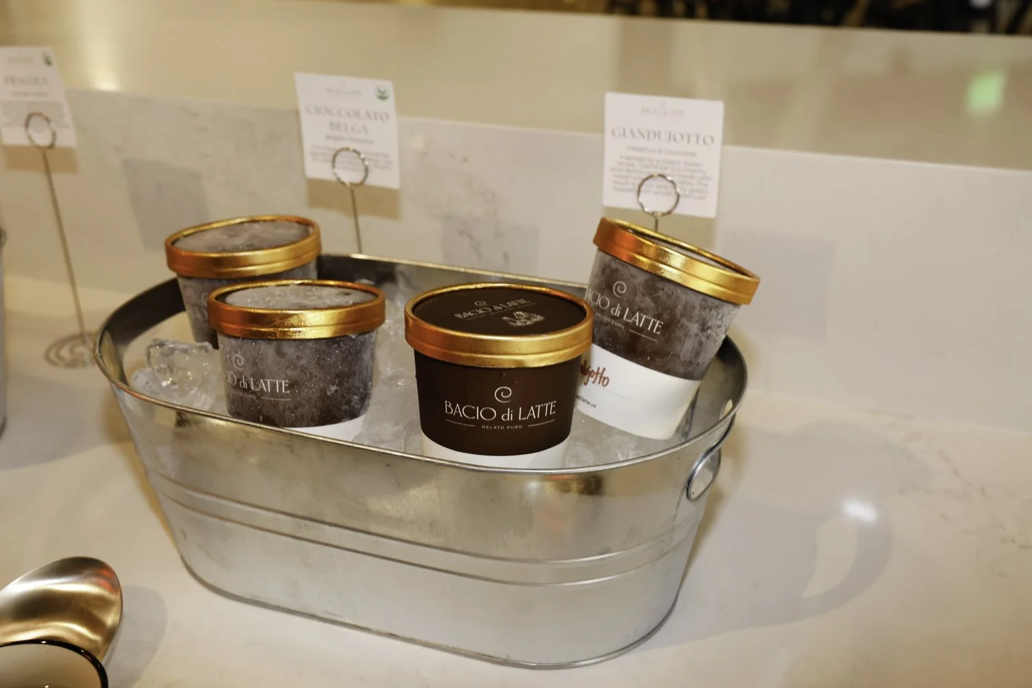 Four small cups of gelato in a metal container filled with ice and labeled with tags behind. The cups have a dark exterior with gold lids and are branded with 'Bacio di Latte' Brex headquarter in San Francisco, CA.