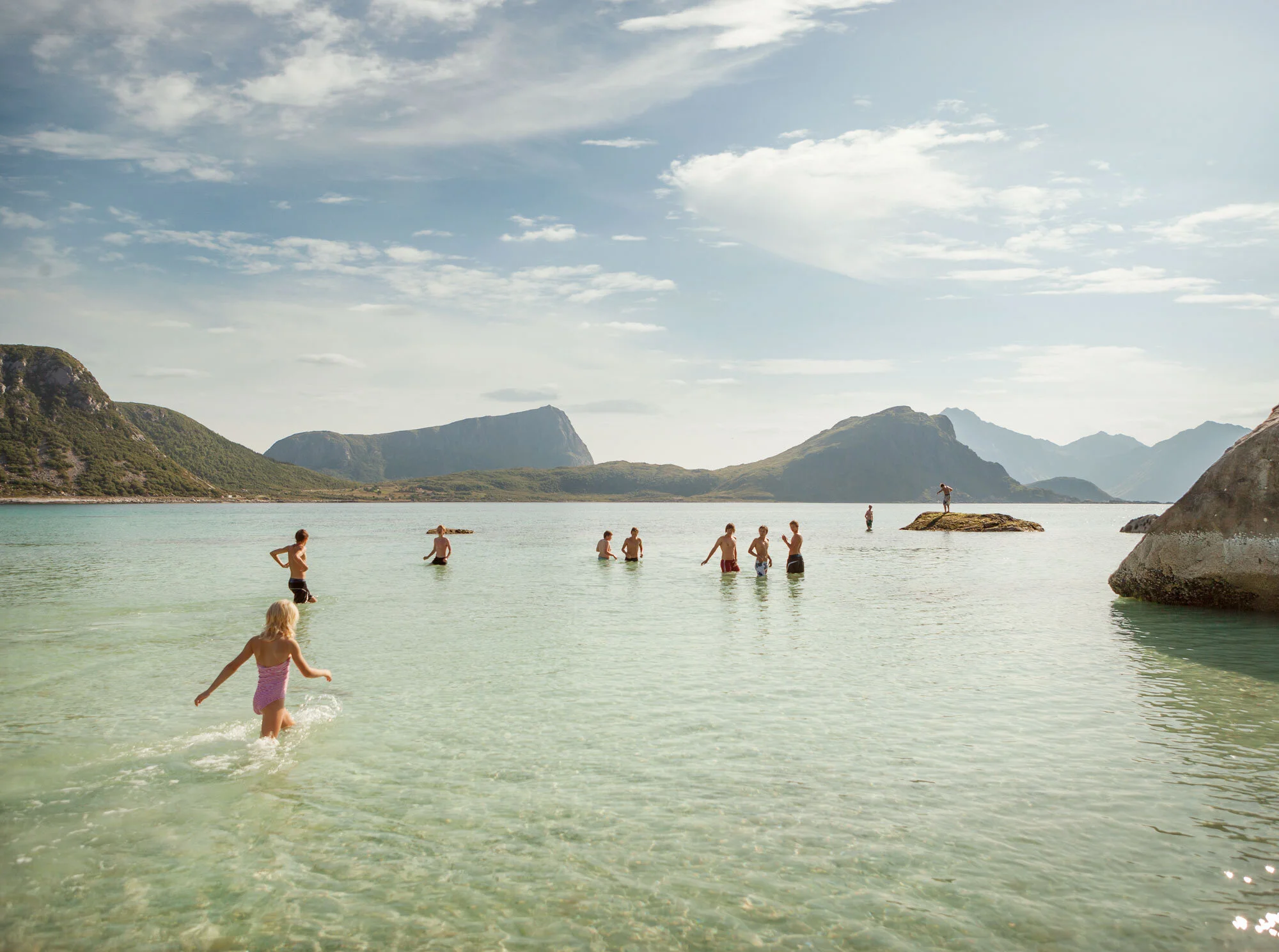 Kids swimming and wading in clear water at a scenic beach in summer with mountains and a partly cloudy sky in the background in Lofoten, Norway.