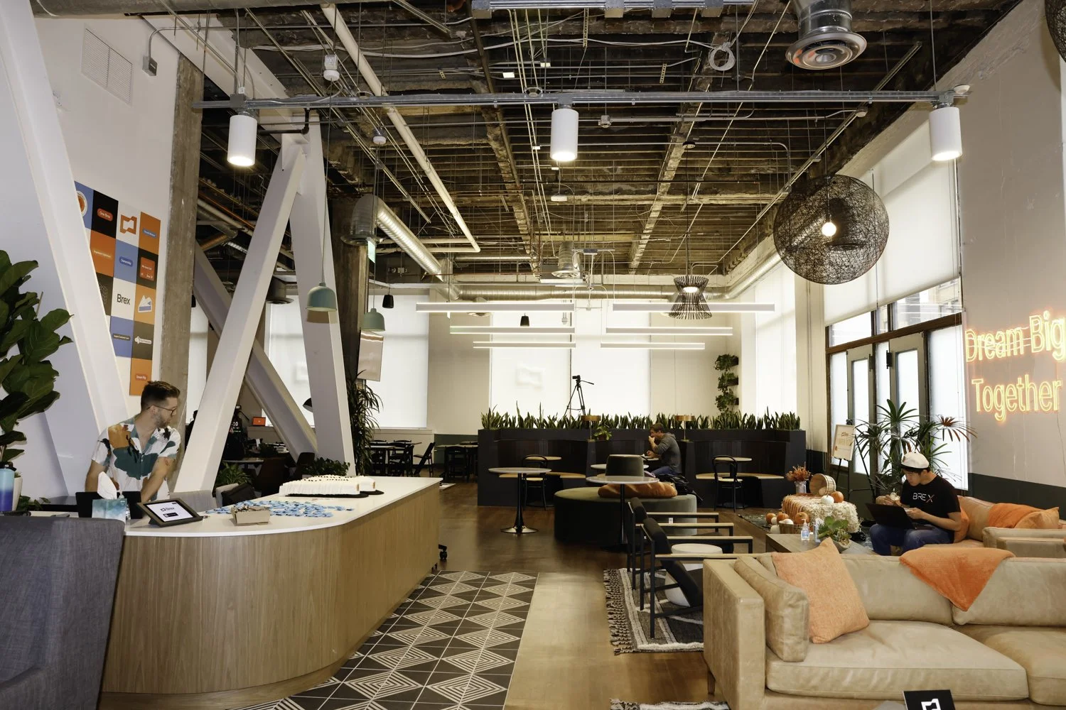 An interior view of a modern, stylish coworking space with exposed ceiling elements and abundant natural light. The space features a reception desk on the left, seating areas with sofas and chairs, potted plants, and a neon sign that reads 'Dream Big