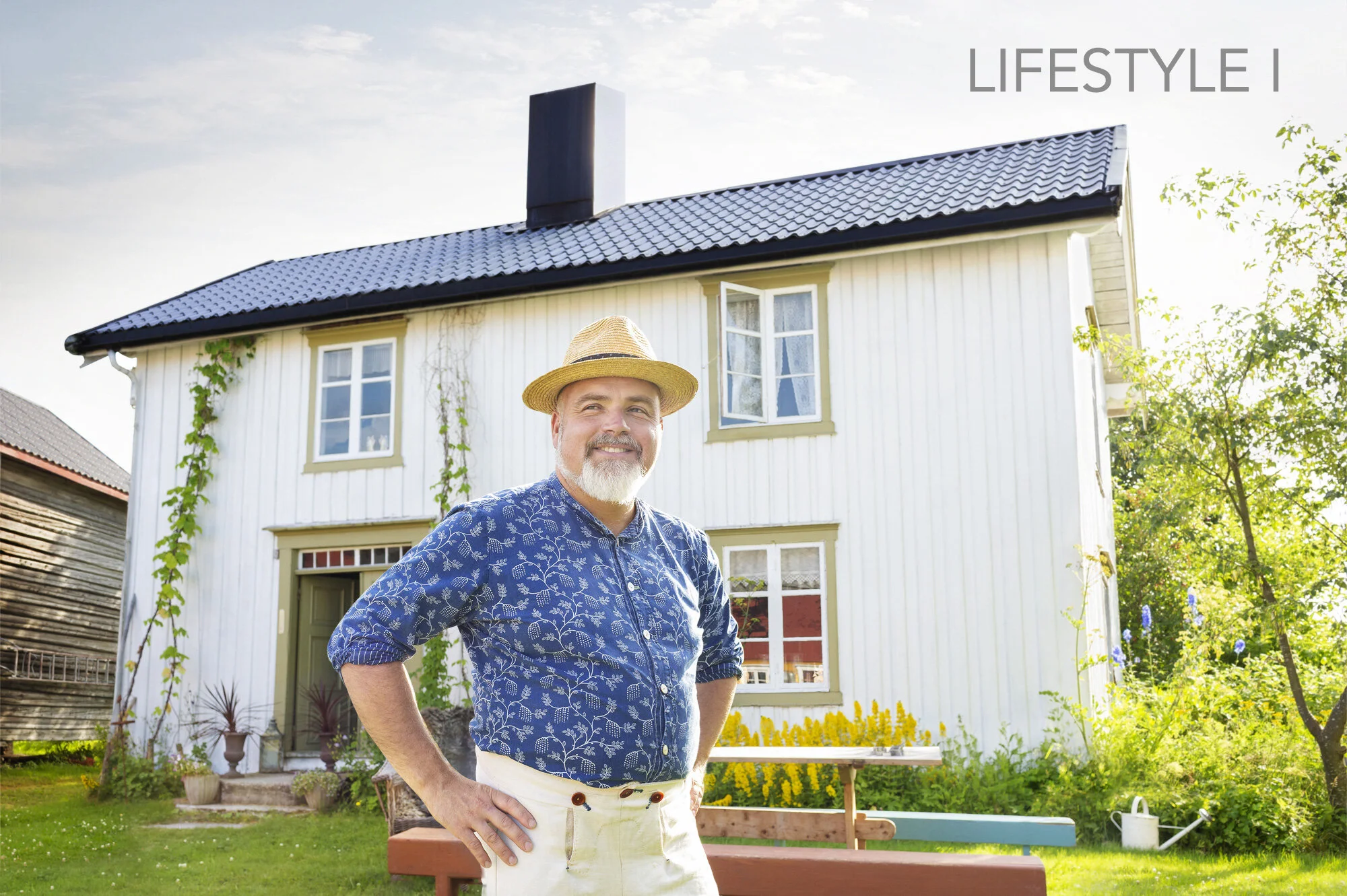 A smiling man with a white beard and mustache, wearing a straw hat and a blue patterned shirt, stands outside in front of a white two-story house with green window frames. The house has a black roof, and there are plants and flowers in the yard, with