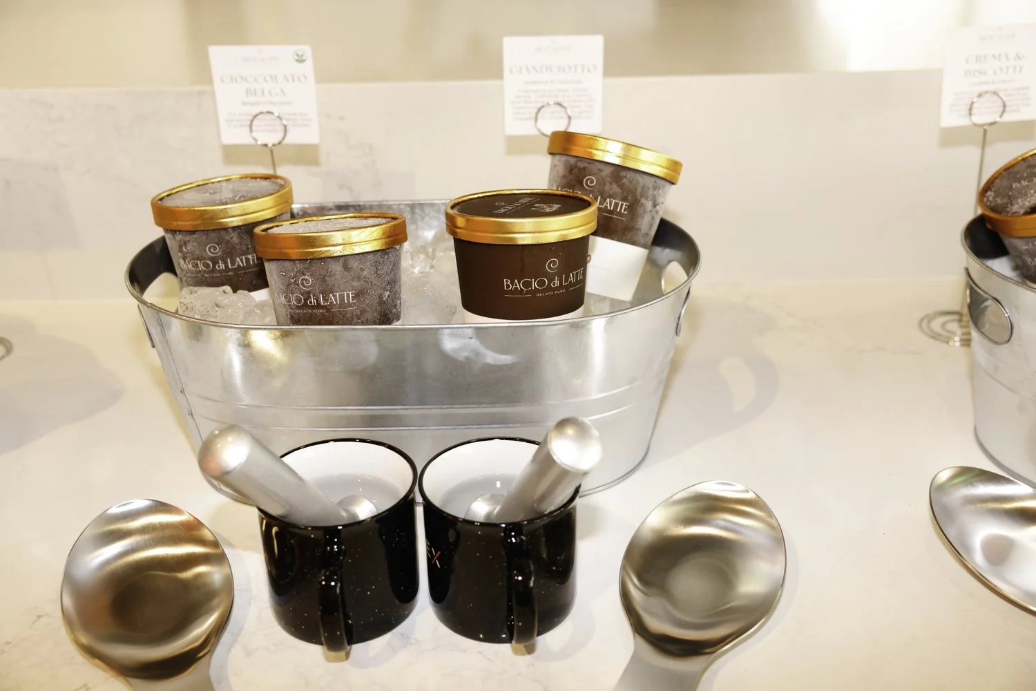 Ice bucket with assorted gelato cups, two black cups with spoons, and metal serving utensils on a white counter at Brex headquarter in San Francisco, CA.