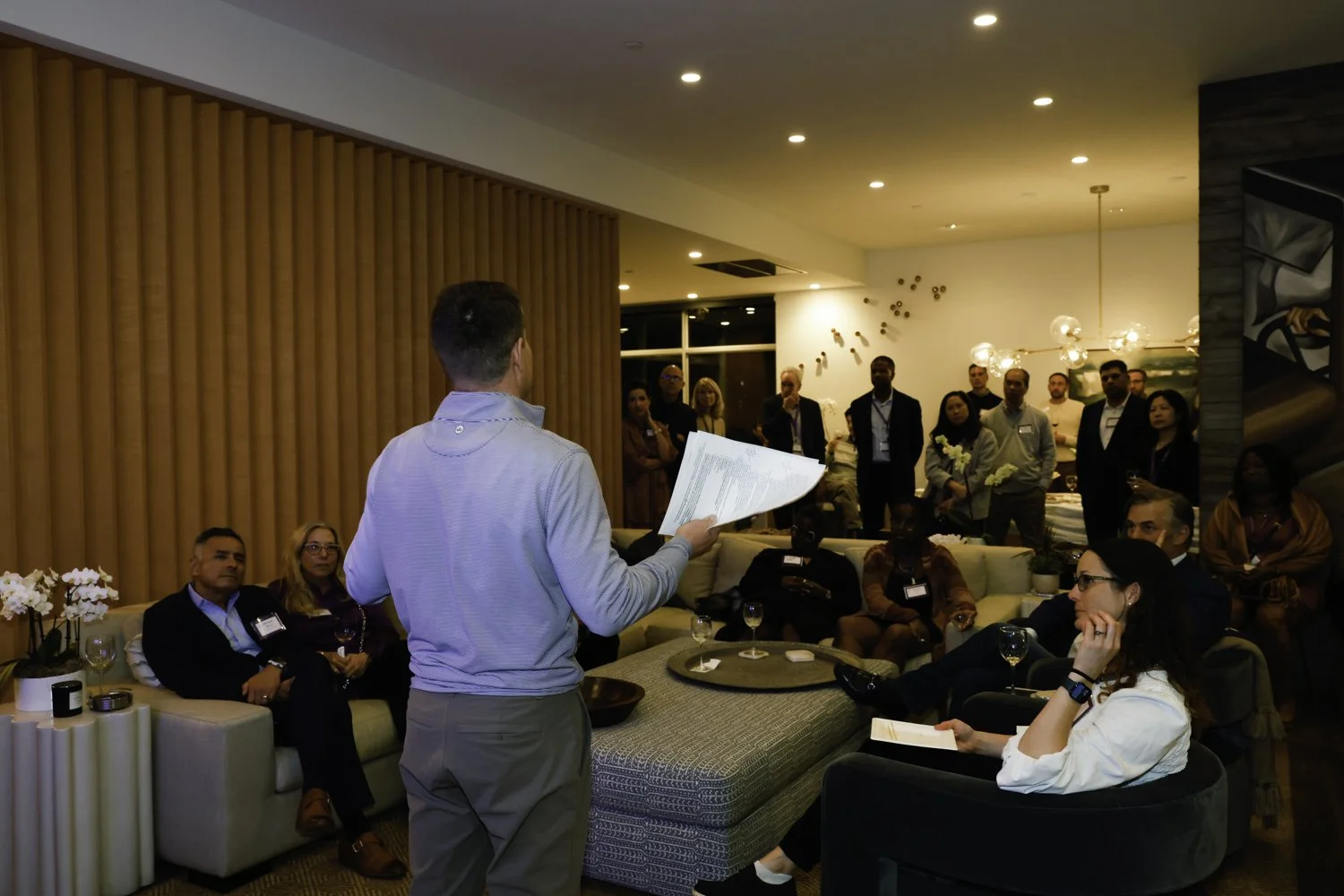 Man in light blue shirt giving a presentation to a group of people seated and standing in a modern living room or lounge area at Northwestern fundraiser in San Francisco, CA.