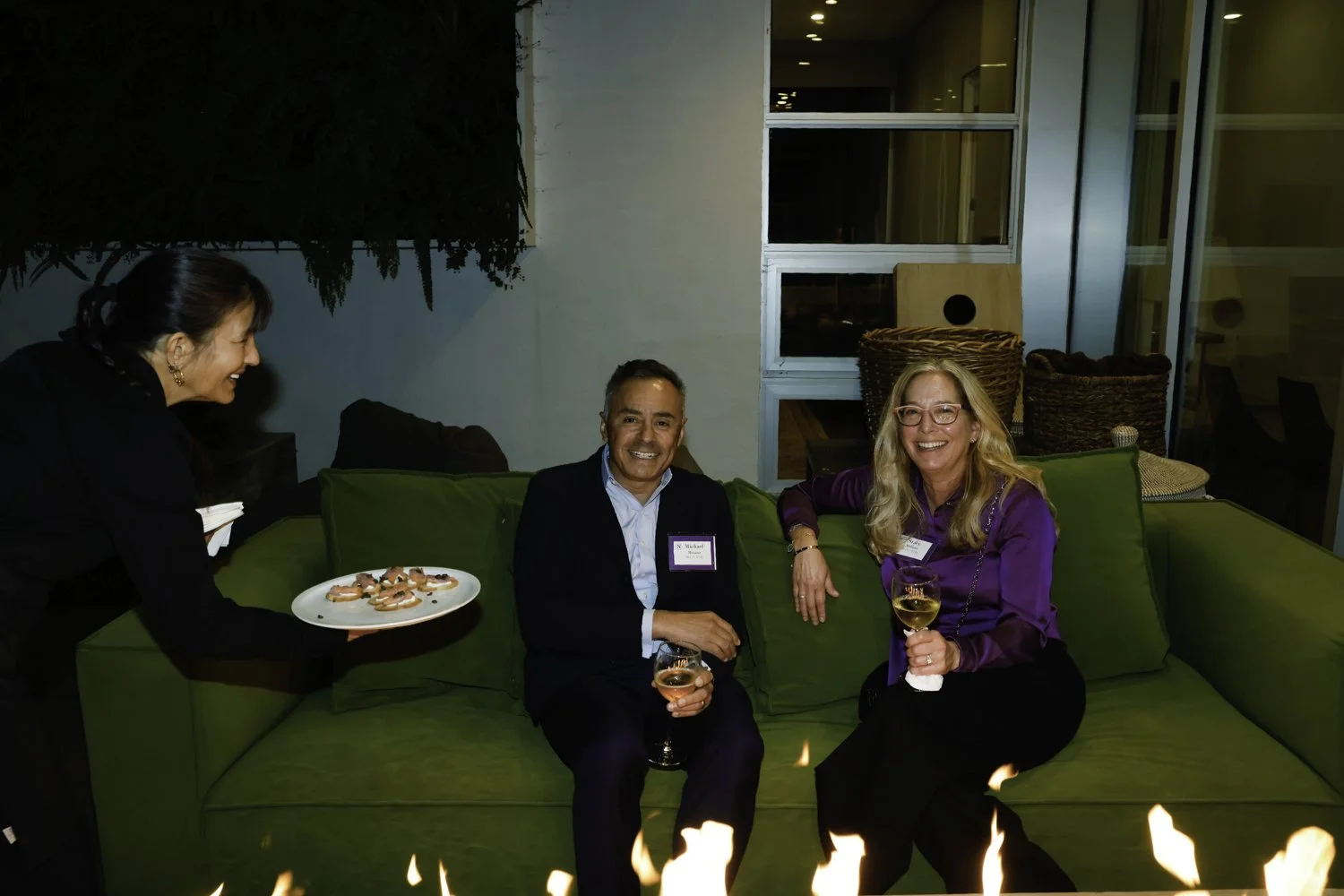 Two men and a woman sitting on a green couch at a social gathering, with a server presenting a plate of appetizers, of the drinks is a glass of white wine, at night in an indoor/outdoor at Northwestern fundraiser in San Francisco, CA.