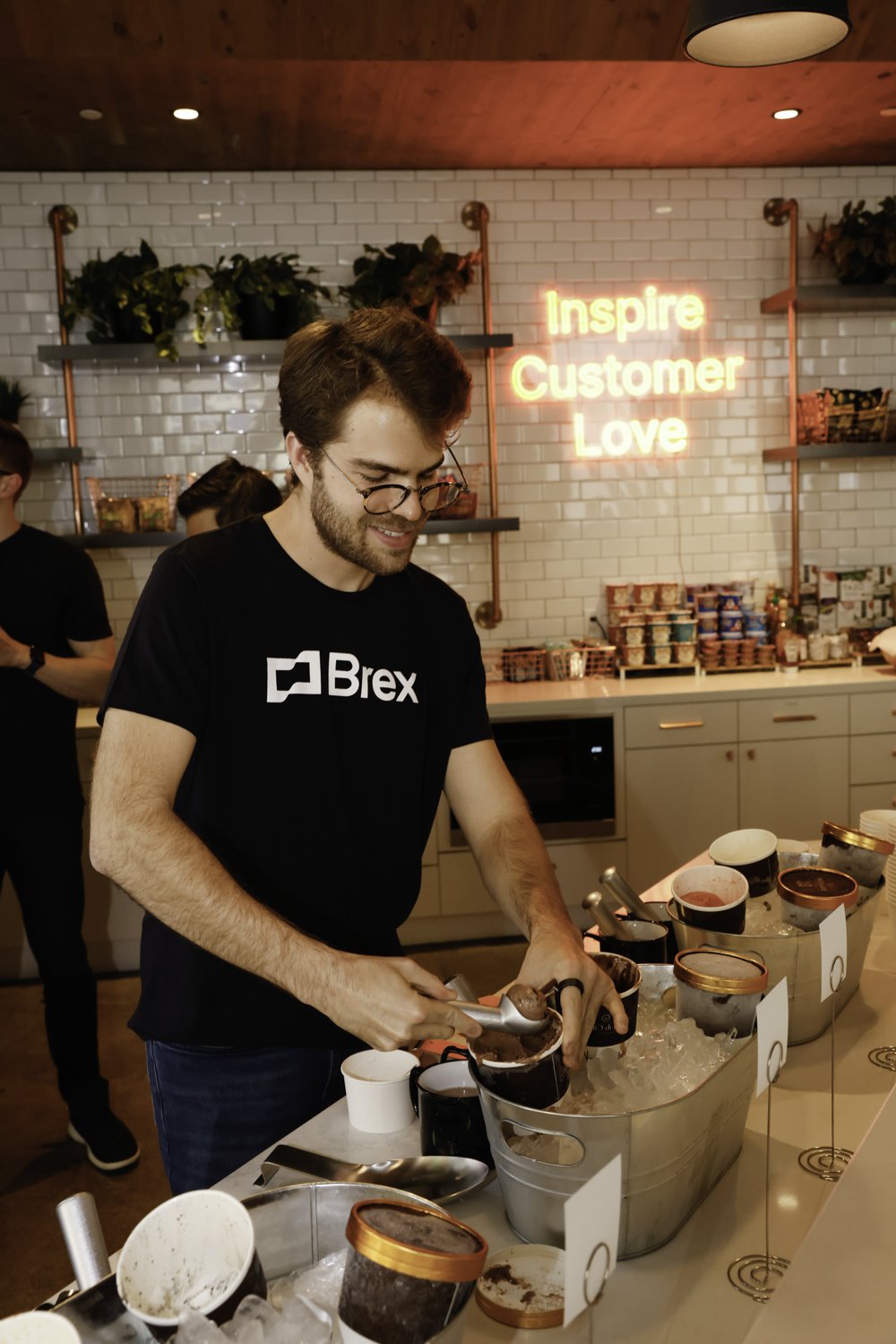 Brex founder with glasses and a beard serving ice cream from containers on a countertop in a modern shop. There are shelves with products and a neon sign that says "Inspire Customer Love" in the background Brex headquarter in San Francisco, CA.