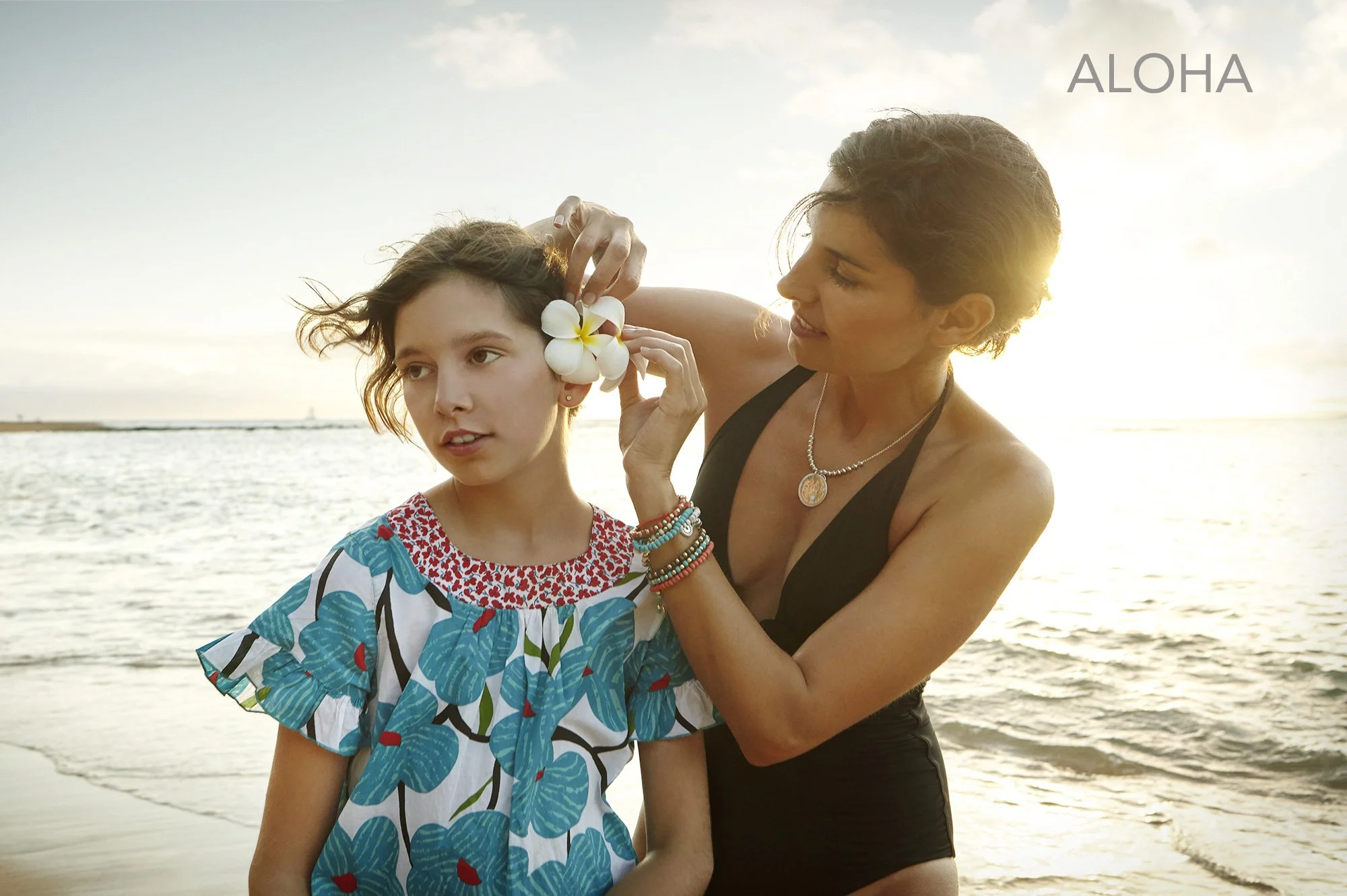 A Latina mother is placing a white flower behind a young girl’s ear on the beach at sunset, with the ocean in the background in Maui, Hawai'i. The girl is wearing a blue and white flower patterned dress and the mother is wearing a black swimsuit.