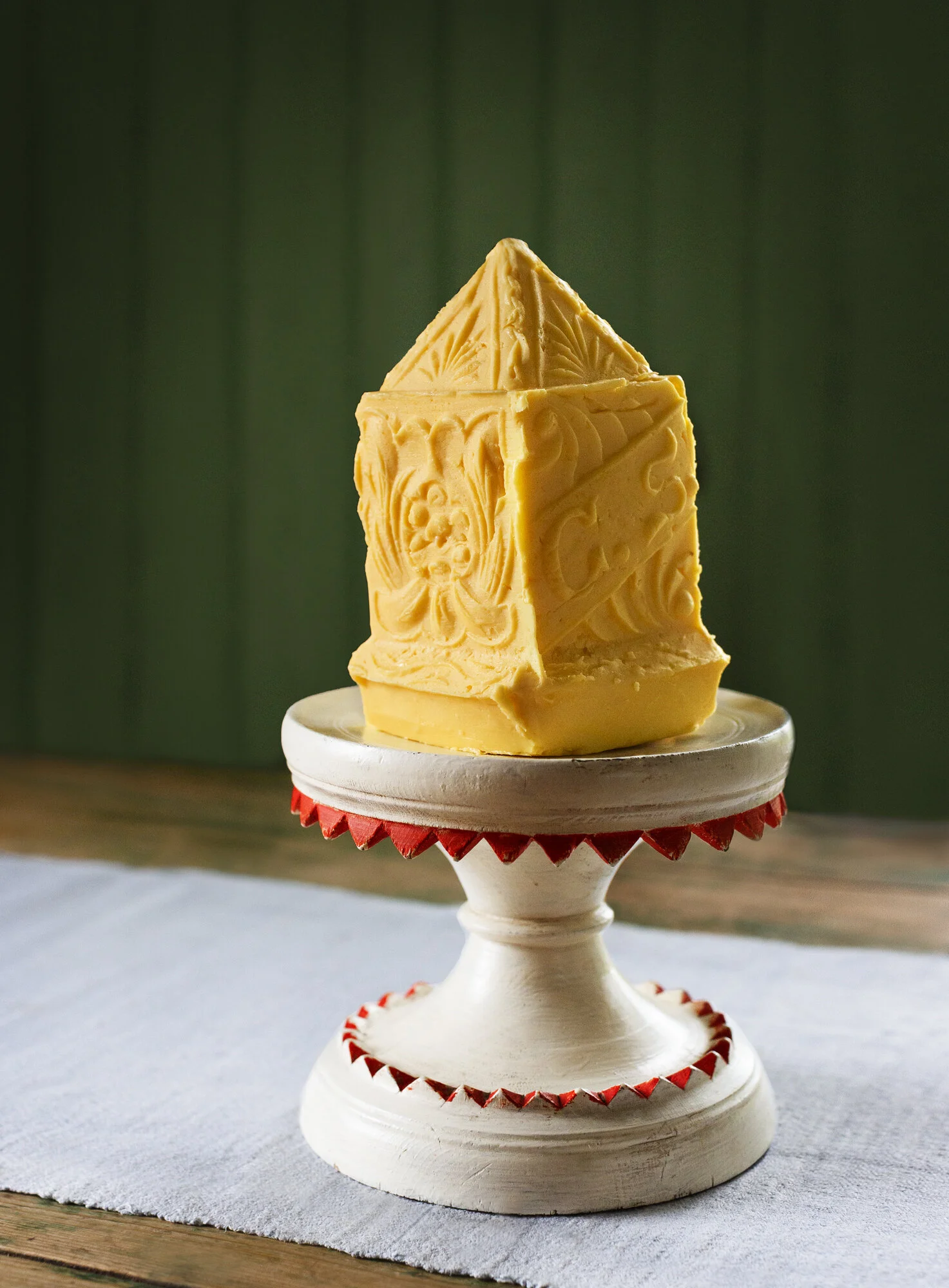 A decorative yellow cheese wedge with intricate carvings, placed on a white pedestal with red accents, on a white tablecloth against a green wooden background.