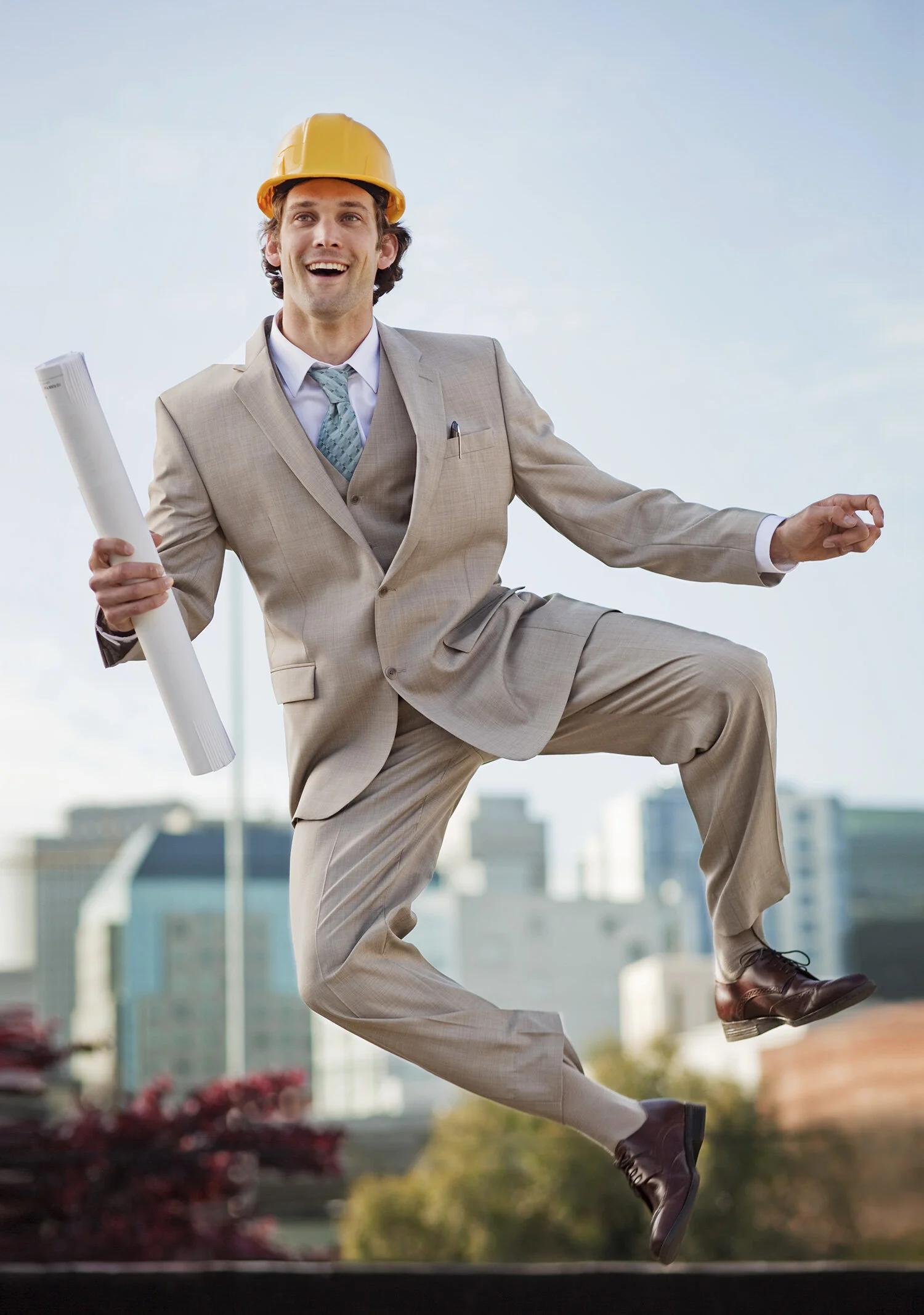 A man in a beige business suit and yellow construction helmet jumping in the air outdoors, holding rolled-up blueprints, with a cityscape background in San Francisco, CA.