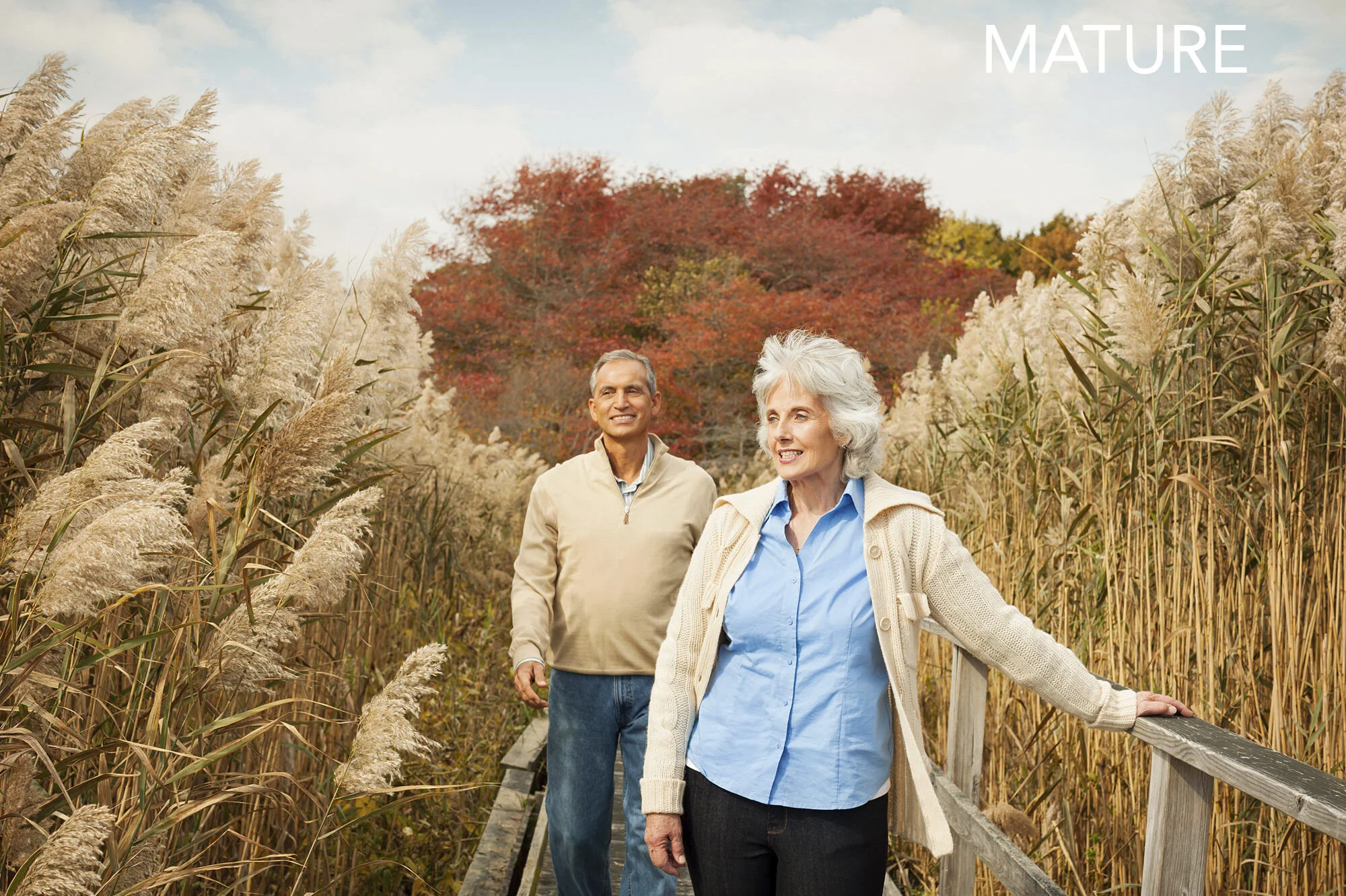 Elderly woman and an elderly man walking on a wooden trail surrounded by tall beige grass with autumn trees in the background on the coast in New Jersey. The woman is smiling and resting her hand on the railing, while the man is smiling behind her. 