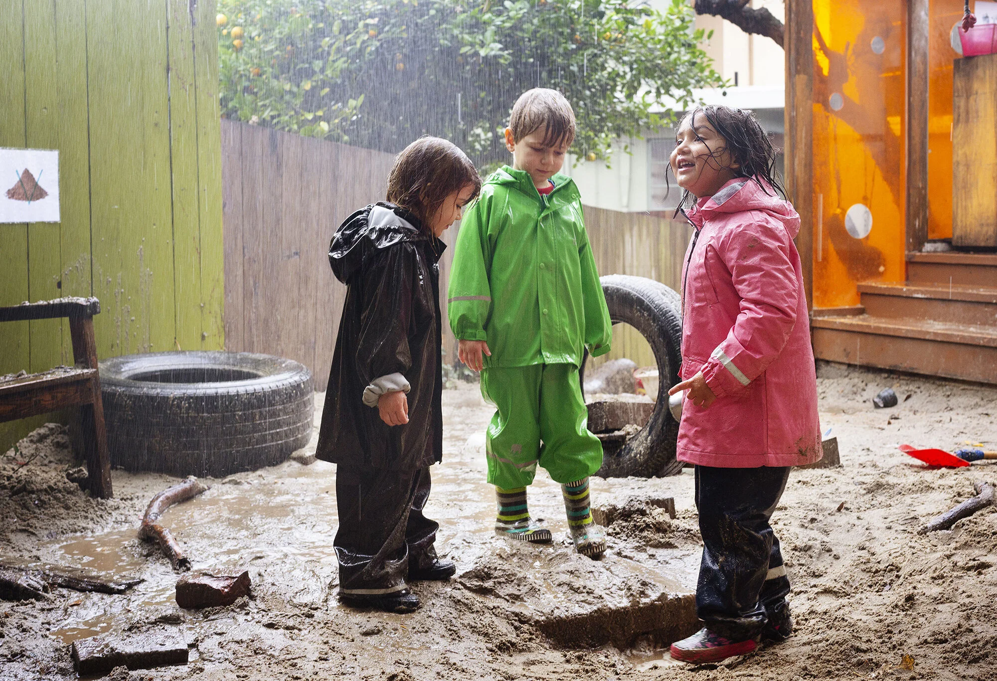 Three children in colorful raincoats and rainboots playing in a muddy sandbox at The Scandinavian School in San Francisco, CA,  during rainy weather. 