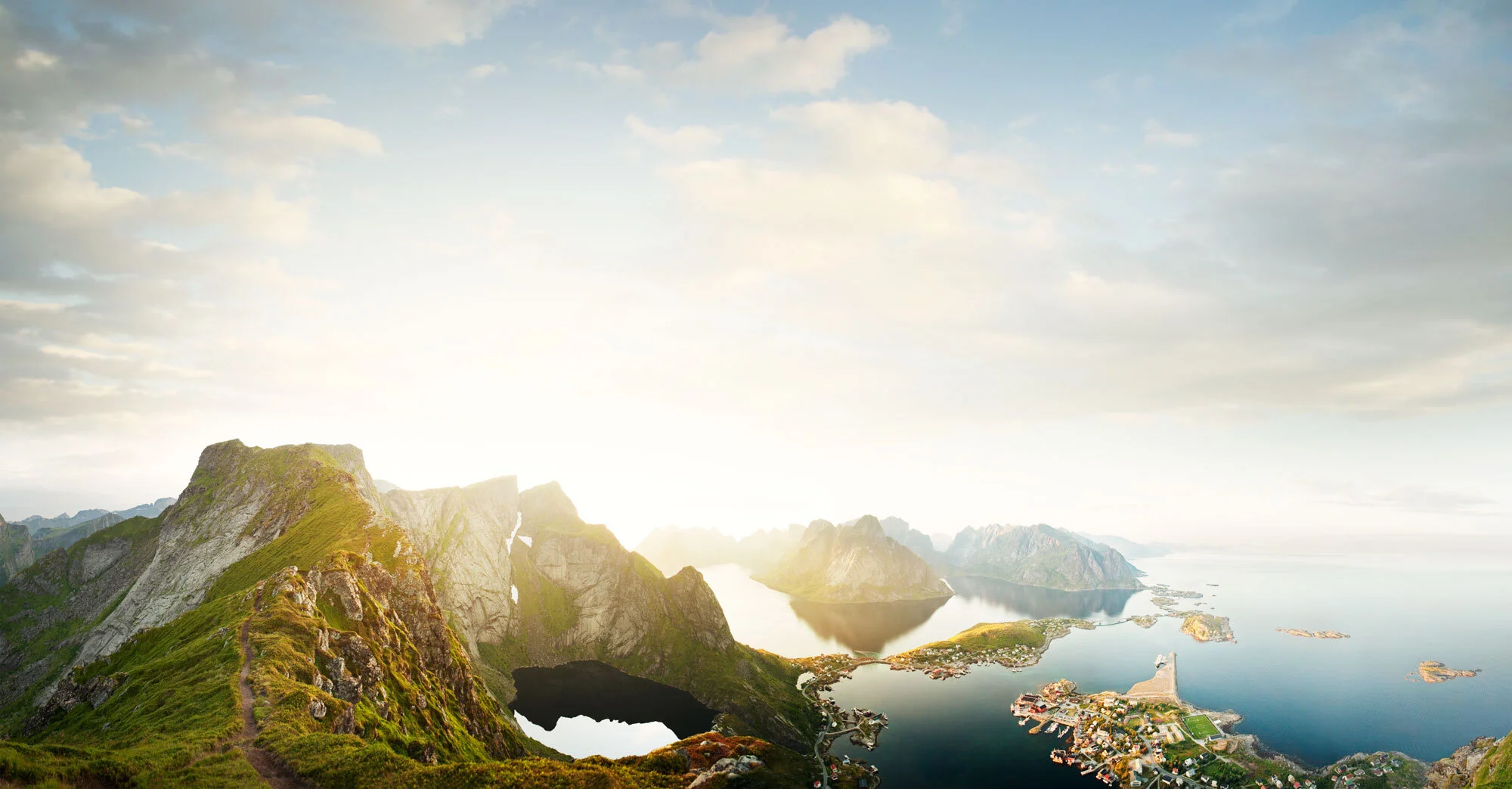 Panoramic and aerial view of a coastal landscape with mountains, a fjord, and a small town, during sunrise or sunset in Lofoten, Norway.