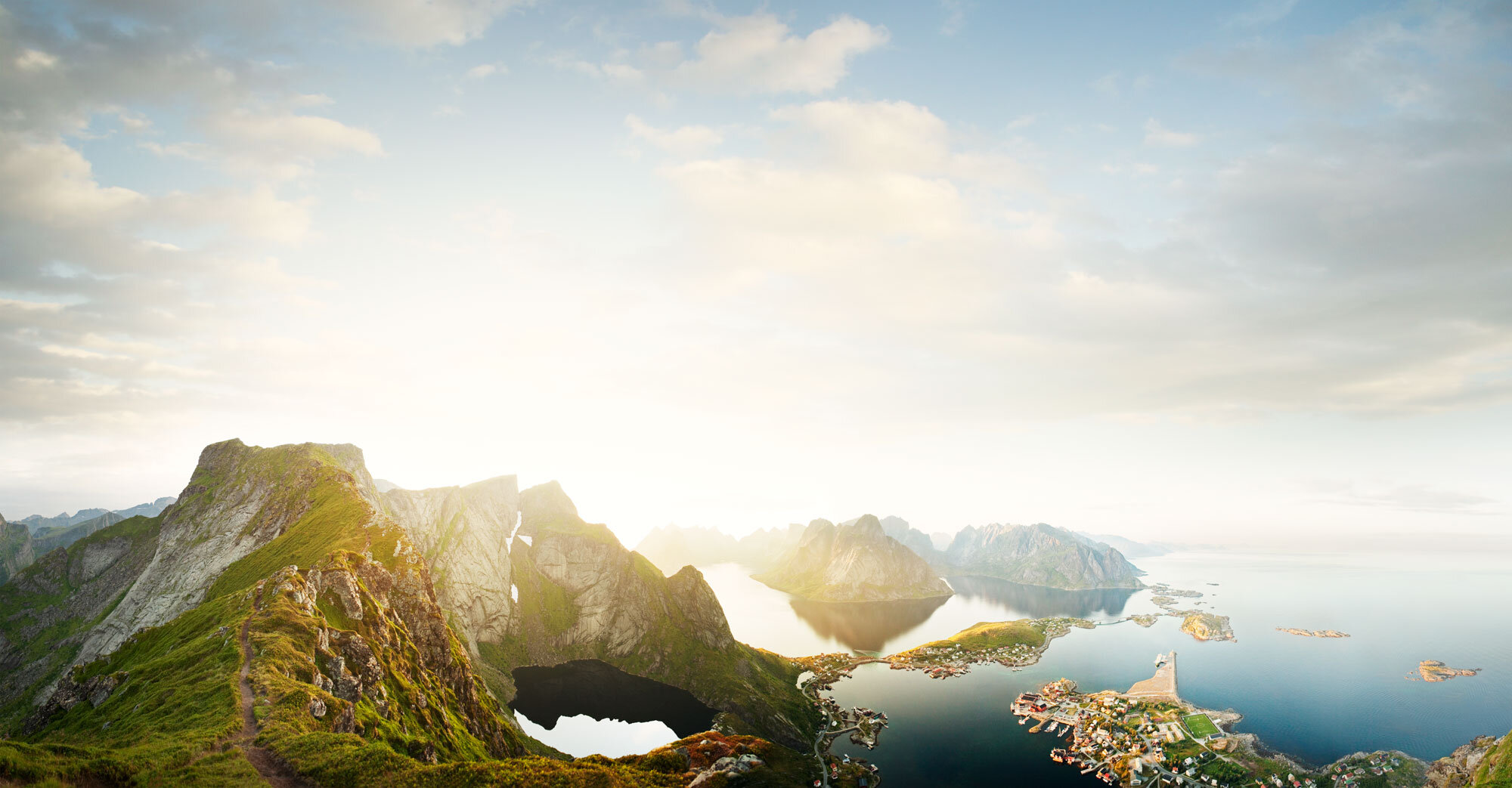 Panoramic and aerial view of a coastal landscape with mountains, a fjord, and a small town, during sunrise or sunset in Lofoten, Norway.