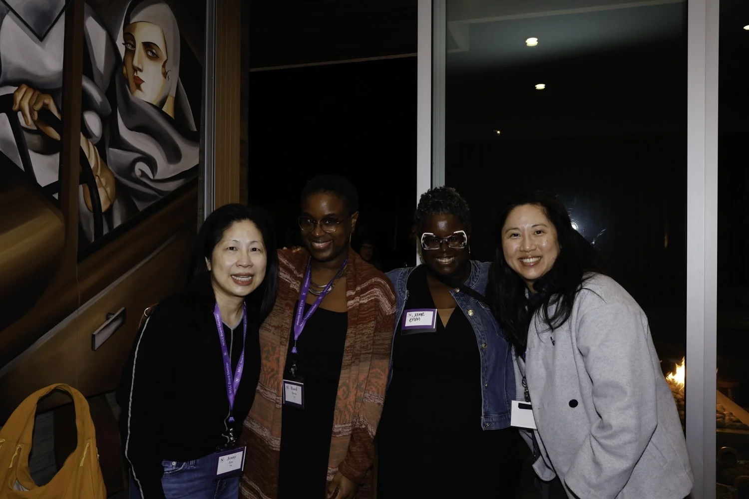 Four women smiling at a social event, standing indoors near a glass wall with a colorful mural of a woman with a scarf, wearing casual attire, and each wearing a name tag with purple lanyards at Northwestern fundraiser in San Francisco, CA.