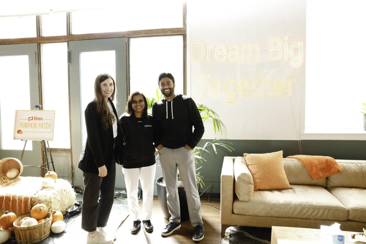 Three people standing together inside a room with autumn decorations and a motivational sign. The left person is a woman with long brown hair wearing a black blazer and white shoes. The middle person is a woman with shoulder-length hair wearing a bla