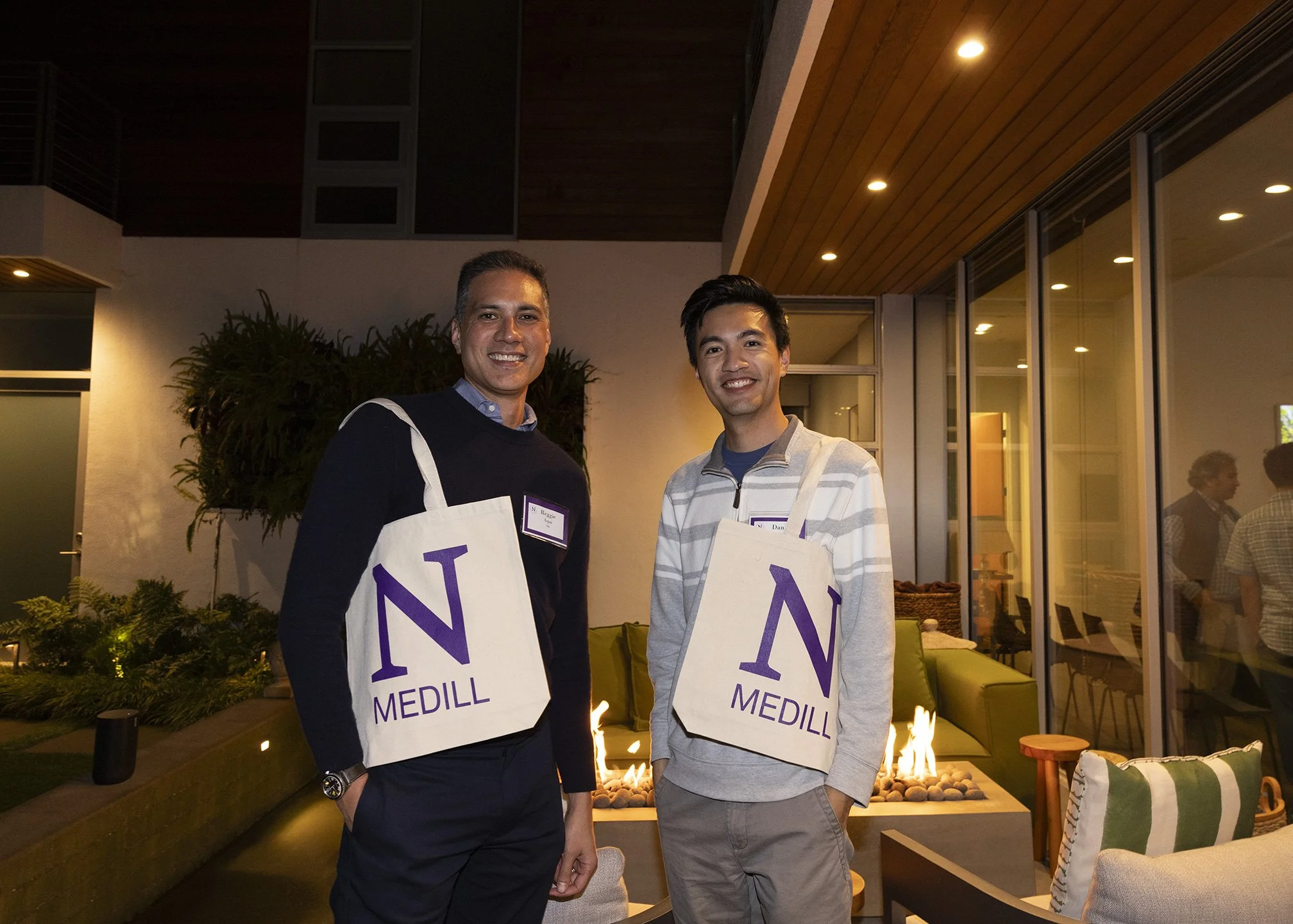 Reggie Aqui from  ABC7 News Bay Area and Dan Hu, Director of Partnerships from CalMatters, standing outside on a patio at night with a Northwestern Medill tote bag over their shoulders, with glass windows in background with reflections of people. Pho