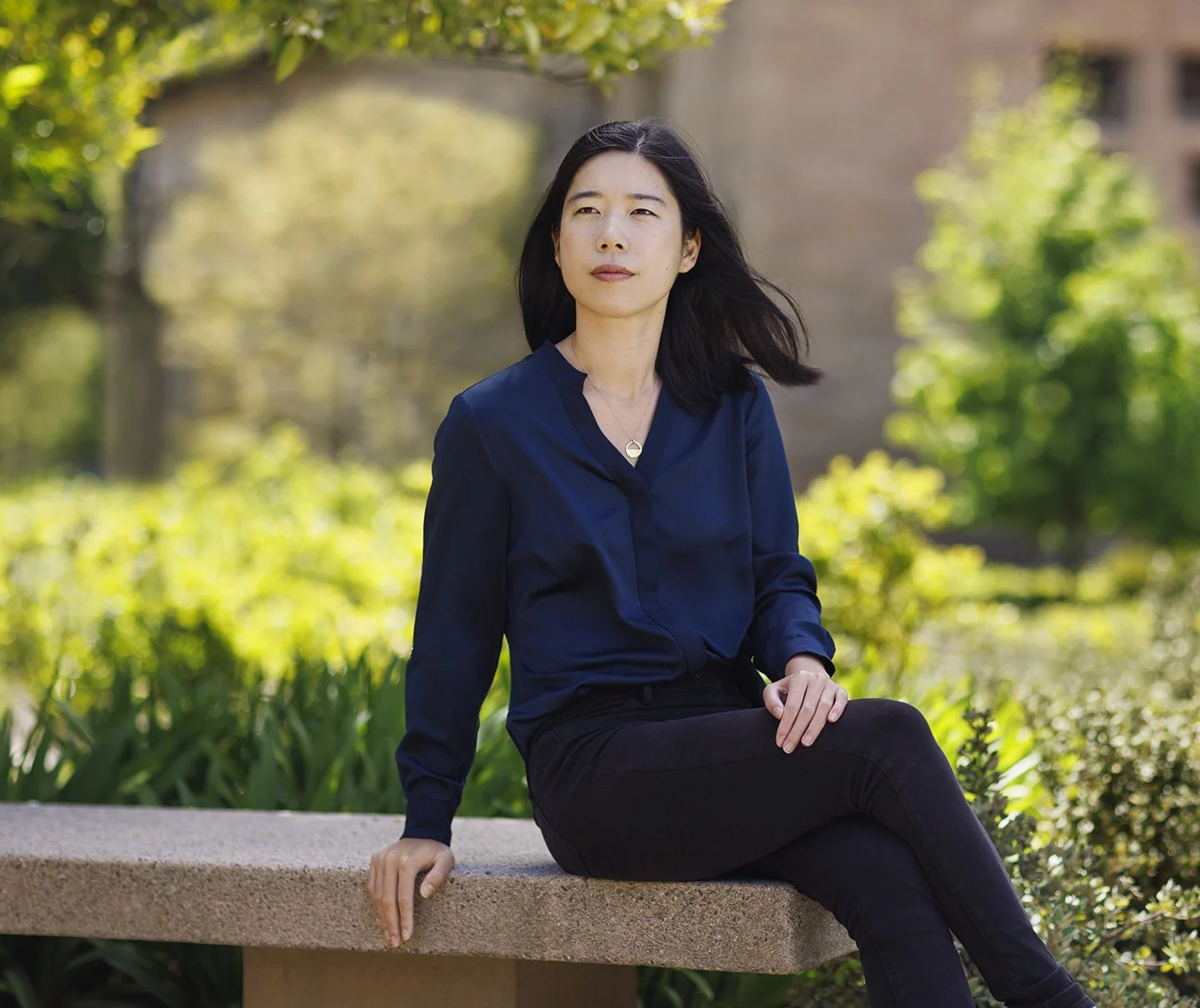 An Asian professor at Stanford University with long black hair is sitting outdoors at a bench with trees and greenery in the background at Stanford University Campus, Stanford, CA.