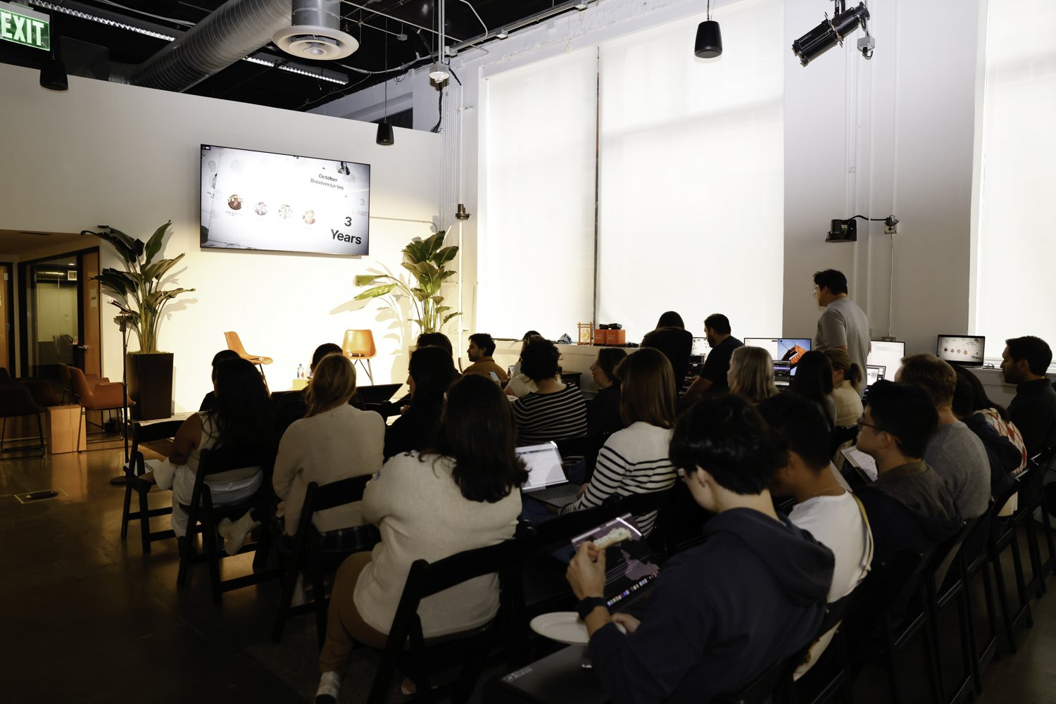 A group of people attending a presentation or conference in a modern, well-lit room with large windows and white walls. There is a large screen displaying a slide, and the audience is seated and listening attentively Brex headquarter in San Francisco