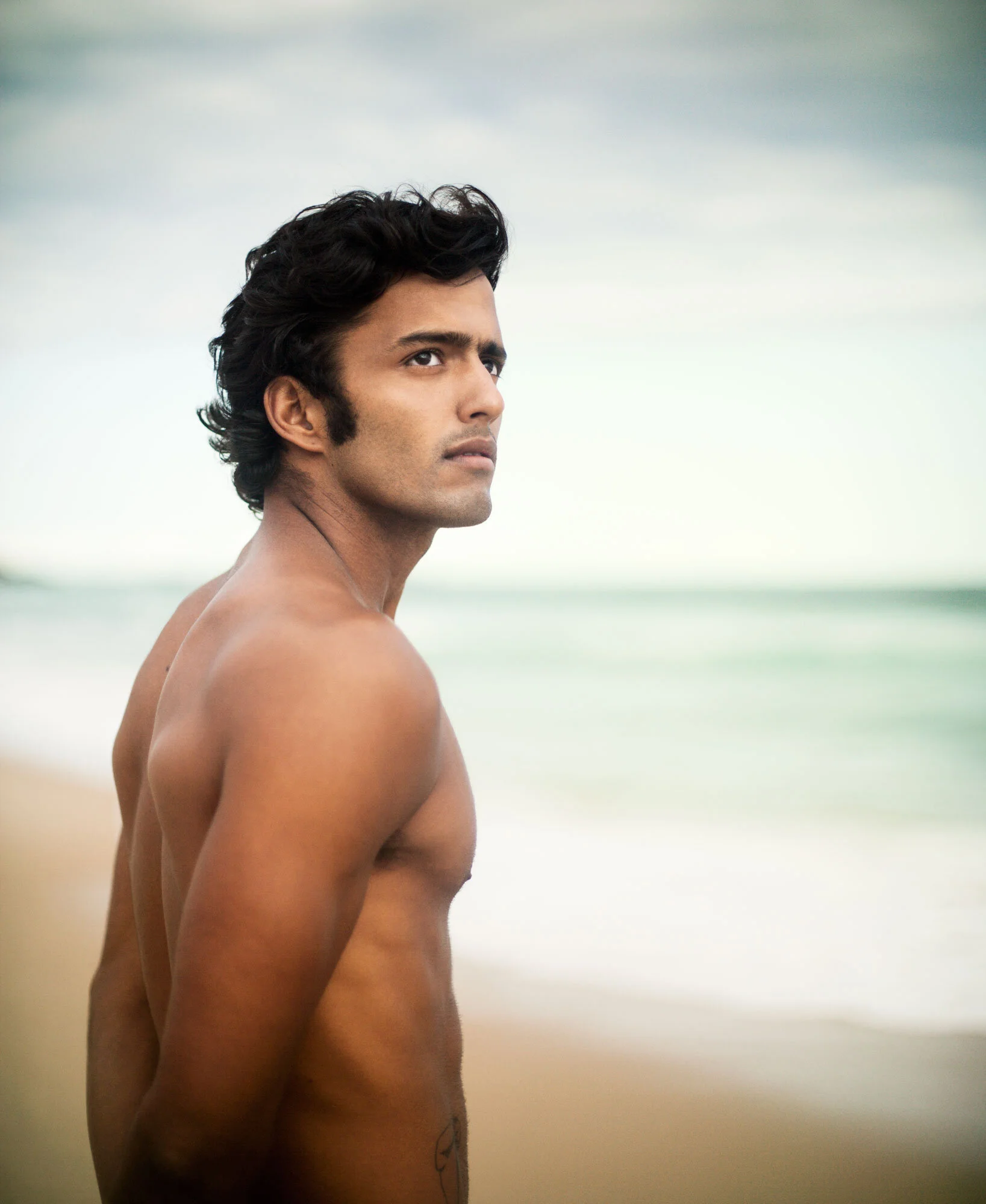 A shirtless man with dark, wavy hair standing on a beach, looking over his shoulder at the camera, with ocean waves and a cloudy sky in the background.