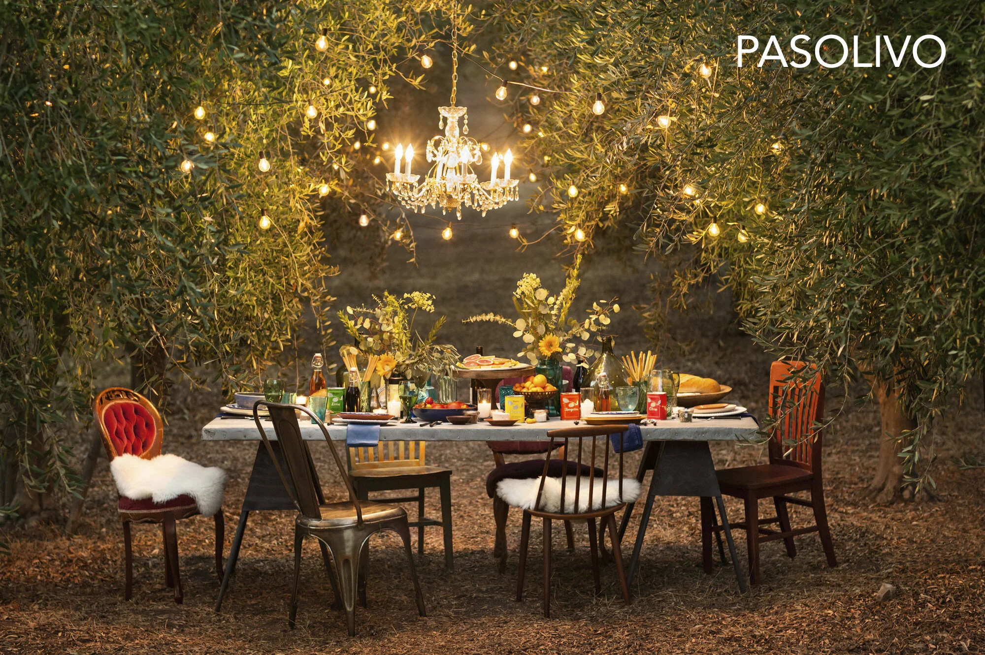 An outdoor dinner table set under trees with string lights and a chandelier, decorated with flowers, food, drinks, and dishes, for a festive gathering in Paso Robles, CA.